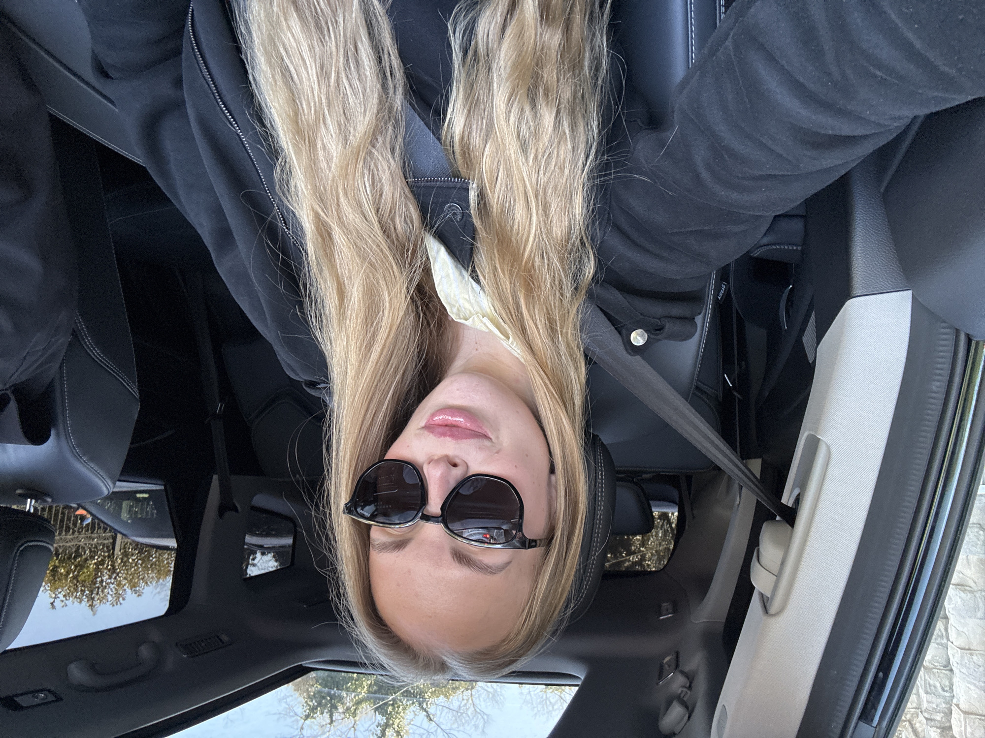 The primary image is an inverted selfie capturing a young woman with long, flowing blonde hair, seated inside a car, likely in Sochi, Russia. She is wearing a dark, possibly black, jacket or hoodie with a visible zipper and a silver snap button detail. Her face is partially obscured by large, round-framed dark sunglasses, but her lower face shows glossy pink lips.
The car's interior is visible, featuring black leather seats with white stitching and black headrests. A black seatbelt is buckled across her chest, securing her in the vehicle. The interior also includes a grey A-pillar with a handle, a grey roof lining, and a panoramic glass sunroof. Through the sunroof and side windows, a clear blue sky and the upper parts of trees or foliage with some bare branches are visible, indicating sunny daytime weather. A light-colored, textured wall or building can also be seen outside the car.
Notable details include the text "AIRBAG" visible on the car's side panel near the seatbelt buckle mechanism. The woman's expression, though largely hidden by sunglasses, appears neutral or slightly relaxed. The overall scene suggests a calm moment, possibly while waiting or during a break in travel.