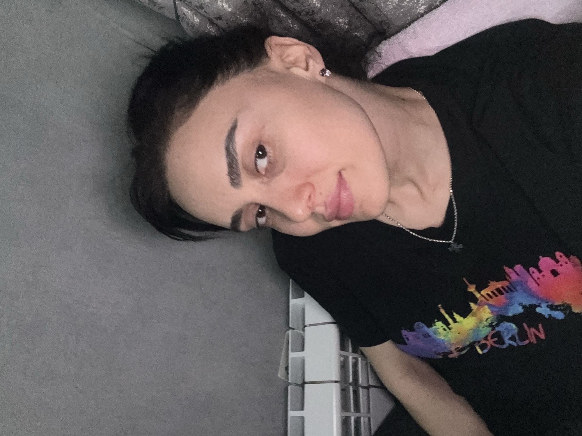 A woman is seen in a close-up shot. She has dark hair, prominent eyebrows, and is wearing an earring. She is also wearing a black t-shirt with a colorful skyline design and the word "BERLIN" printed on it. A silver necklace is visible around her neck. The background includes a gray wall and a white tiled surface. The location in the city of Tbilisi, Georgia is not apparent in the image.