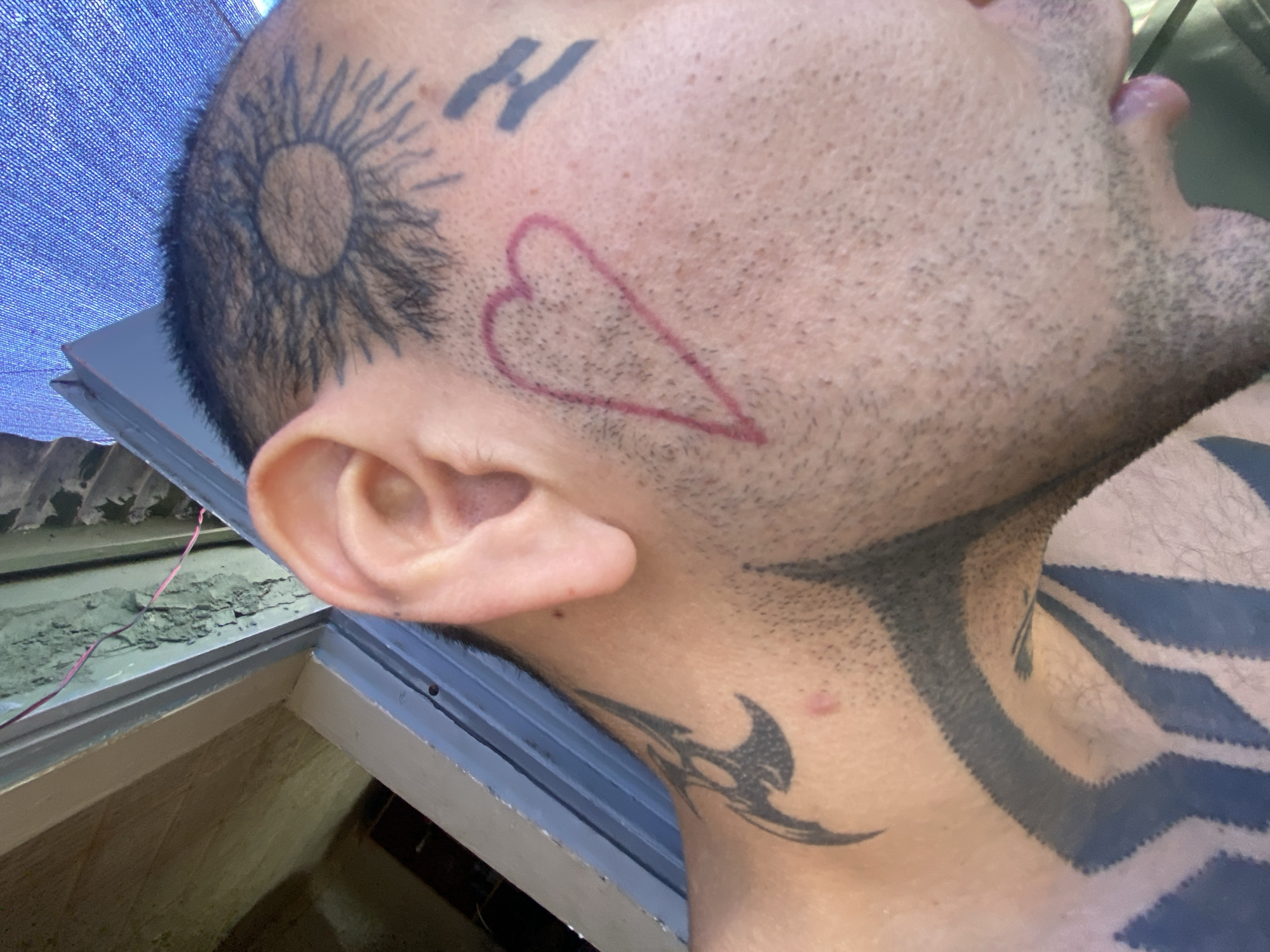 A medium-close-up shot shows the side of a person's head and neck, with a pale skin tone. The person has several tattoos on their head and neck. On the head, there's a tattoo resembling a sun, the letter "H," and an outlined heart. The neck displays a tribal-style tattoo. The person has short, dark hair and facial hair. The background consists of a blue tarp and a window frame, suggesting an indoor environment. The location within the city of Buenos Aires, Argentina cannot be determined with the details visible in the provided image.