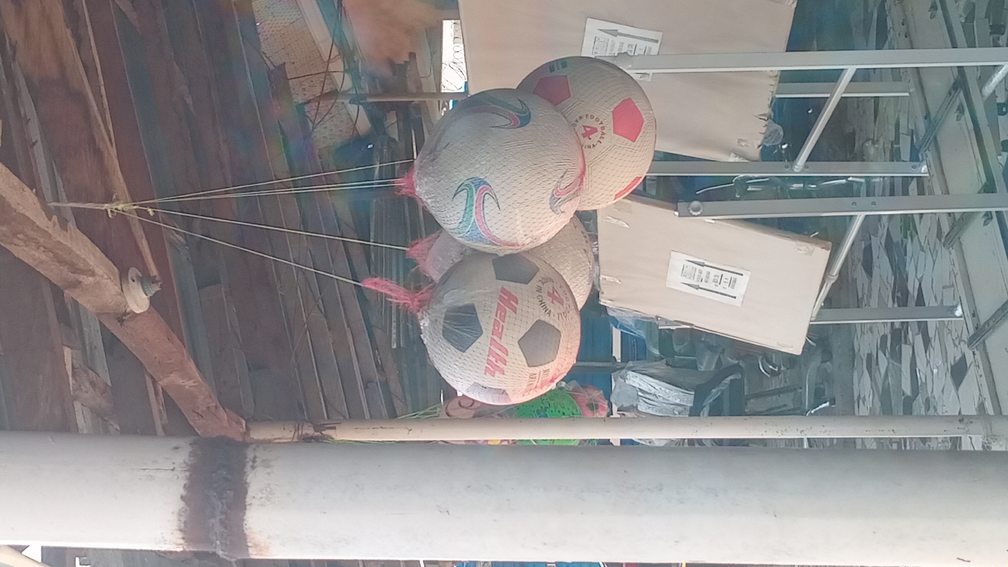 The image depicts a market stall in Jalingo, Nigeria, where several soccer balls are displayed, hanging from the ceiling by strings. The stall has a rustic wooden roof supported by a white column and beams. One soccer ball is prominently in the foreground, wrapped in plastic netting, with the word "Health" visible in red lettering. Other soccer balls, some with colorful patterns, are also visible, some partially obscured. In the background, there are various items, including what appear to be more merchandise covered with cloth or plastic, and metal stands with what look like displays or signs. The ground appears to be paved with irregularly shaped tiles. The overall scene suggests an outdoor market environment with goods for sale.