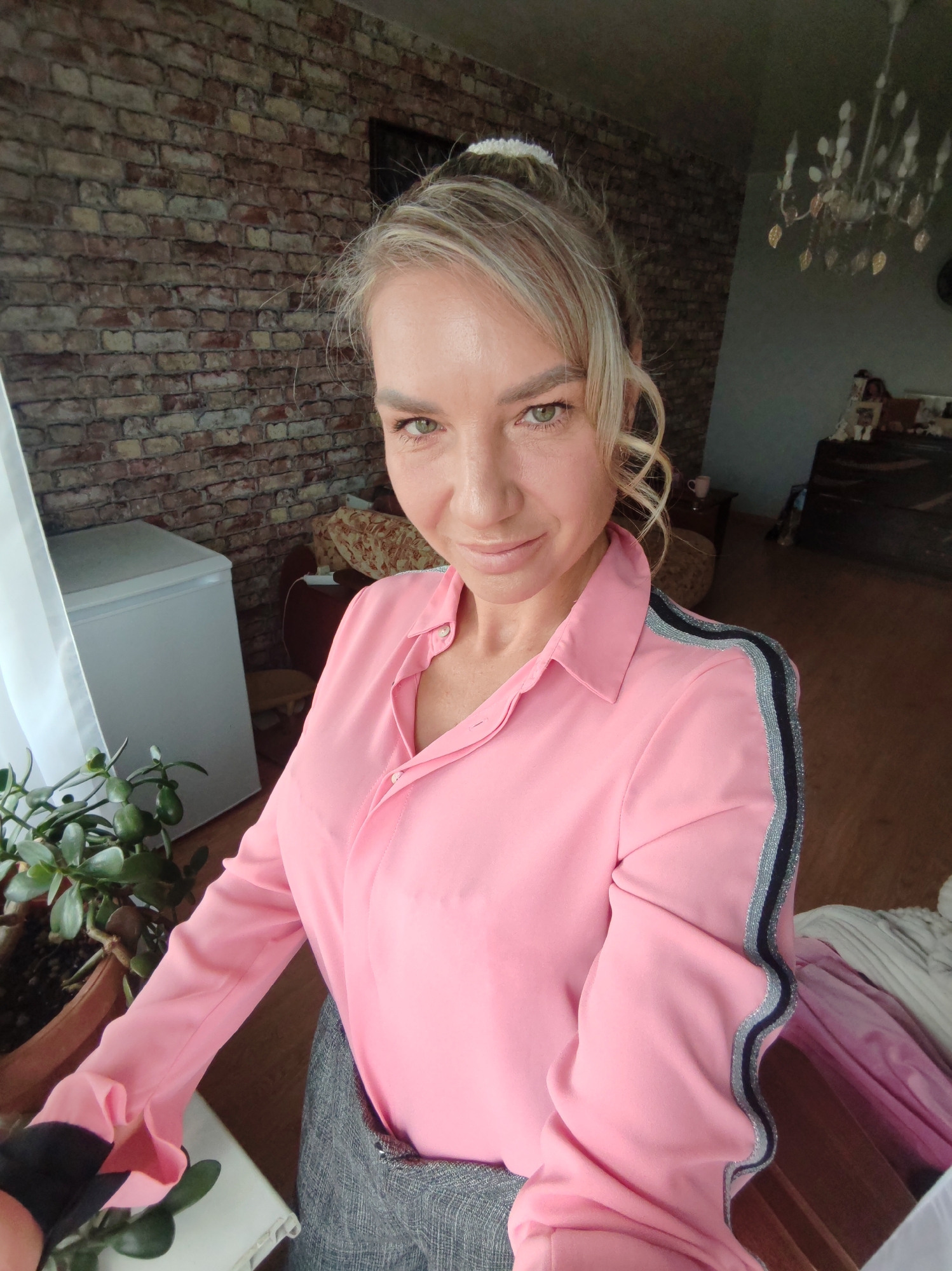 A blonde woman, likely in her 30s or 40s, takes a selfie indoors. She is wearing a pink collared shirt with silver and black stripes on the sleeves, and a grey textured skirt. Her hair is pulled back in a messy bun with some loose curls framing her face. She has green eyes and is smiling slightly.
The background suggests a room in an apartment, possibly in Petropavlovsk-Kamchatsky, Russia, though this cannot be definitively confirmed from the image alone. To her left, a white refrigerator is partially visible next to a window draped with a sheer white curtain. In front of the window, a potted plant with green succulent leaves is placed. The wall behind her is a textured brick pattern. Further into the room, a chandelier is visible hanging from the ceiling. The overall impression is of a casual, domestic setting. The lighting is natural, coming from the window, and illuminates the woman's face.