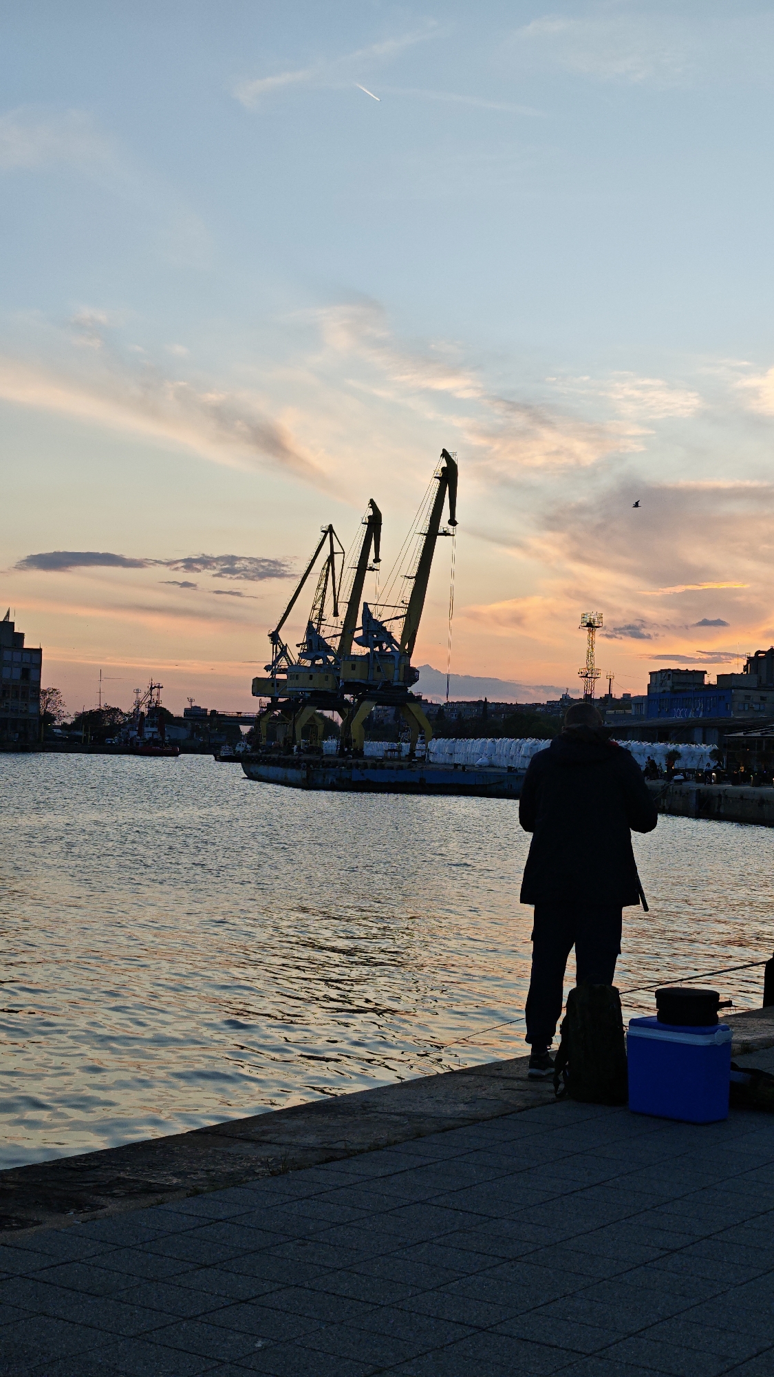 The image shows a scene at the port of Burgas, Bulgaria, during sunset. Three yellow cranes are visible in the background, situated on a barge. In the foreground, a man is standing on a concrete quay, facing the water. He is wearing dark clothing. Next to him are a backpack and a blue cooler. A fishing rod is visible near the edge of the quay. The sky is filled with orange and pink hues from the setting sun, with clouds and a contrail from a jet plane. A bird is flying in the sky. The water reflects the colors of the sunset.