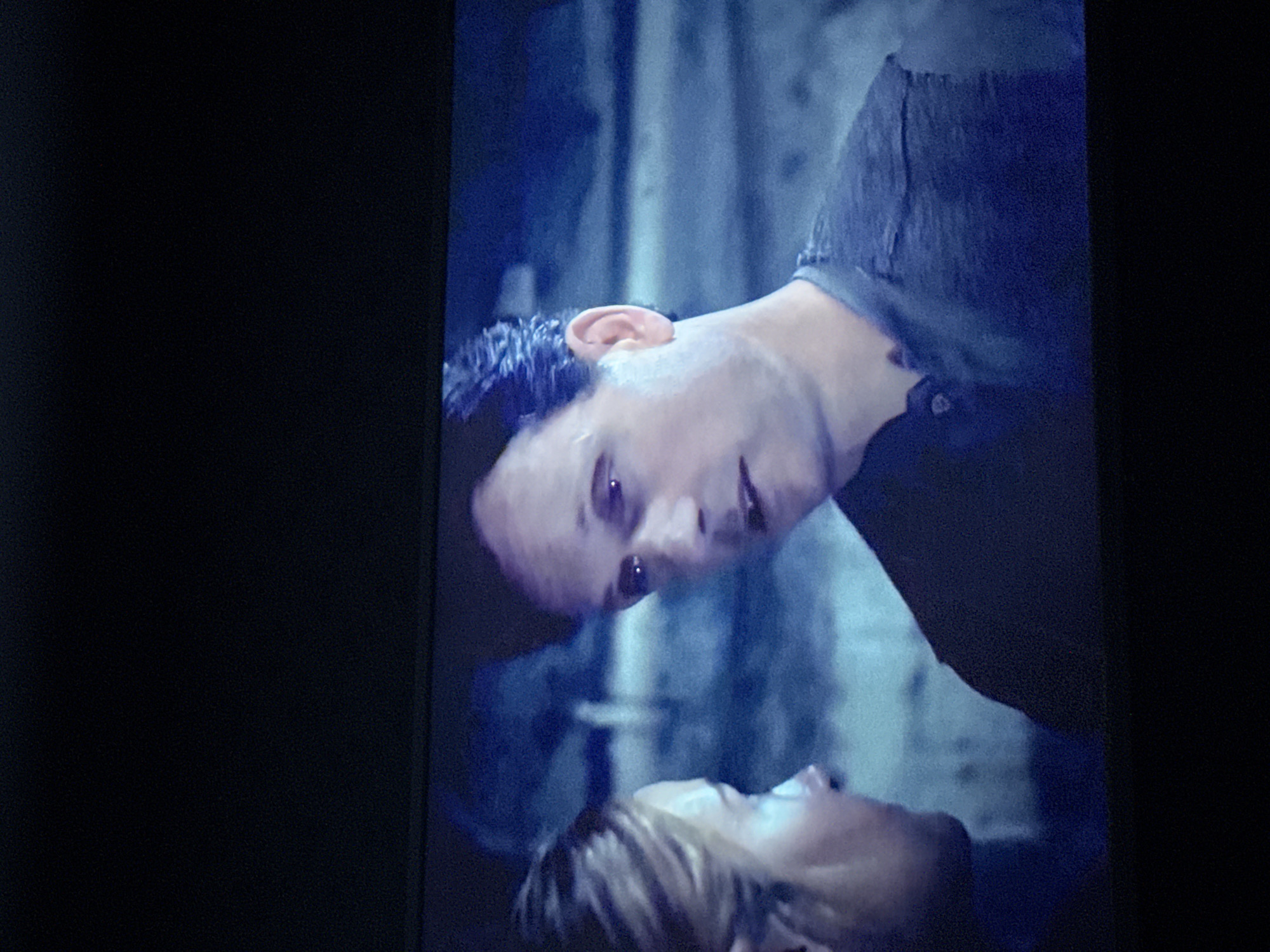 The image shows two people, one man and one woman, in an intimate and intense face-to-face interaction. The man is positioned above the woman, leaning down towards her. Their faces are close, almost touching. The scene is bathed in blue light, obscuring details and giving the image a dramatic, moody atmosphere. The background appears to be an indoor setting, possibly a room with textured walls or draped fabric. The subjects' expressions are ambiguous, potentially indicating emotional tension, intimacy, or conflict. There is no information to suggest that this photograph was taken in Mytishchi, Russia, or any other location.