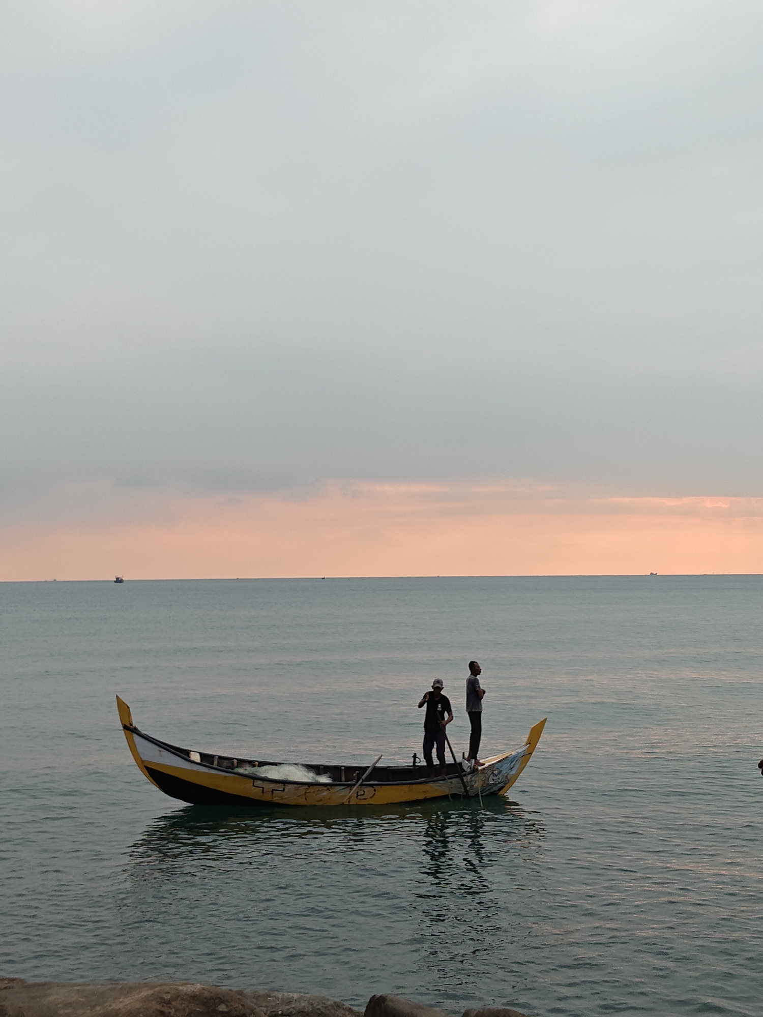 This image captures a coastal scene in Lhokseumawe, Indonesia, at either sunrise or sunset. A light grey sky transitions to a band of soft orange-pink at the horizon, meeting a calm, dark blue-grey ocean. In the mid-ground, a traditional wooden fishing boat, predominantly yellow and black with white trim, floats on the water. A fishing net and oars are visible within the boat. Two adult males are standing inside the vessel. The man on the left, wearing dark clothing and a cap, holds a long pole or oar. The man on the right, dressed in a light-colored t-shirt and dark pants, stands further aft, looking towards the right side of the frame. Multiple dark silhouettes of other vessels are discernible on the distant horizon. The immediate foreground reveals a dark, indistinct rocky or concrete surface.