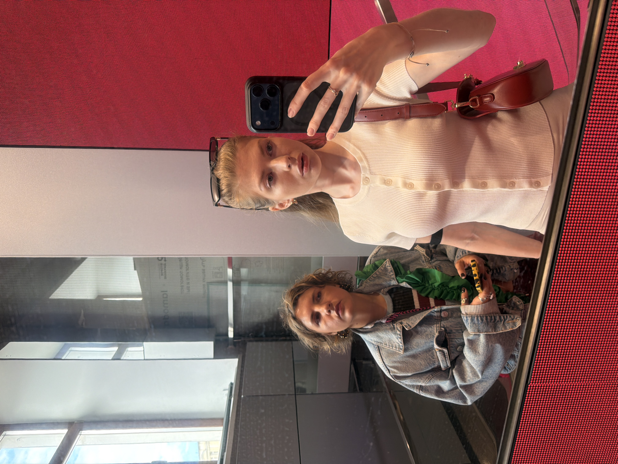 A young woman is taking a selfie in front of a reflective surface in a building. The location is the Bolshaya Dmitrovka Street in Moscow, Russia.
The woman is holding a black smartphone in her right hand, positioned to capture her image in the reflective surface. She is wearing a light-colored ribbed top with button detailing, a small brown bag over her shoulder, and eyeglasses resting on her head. She has blonde hair and is wearing a ring on her ring finger.
The reflection shows another woman wearing a denim jacket and a striped shirt with a green scarf. She is holding a black and yellow object.
The background consists of a wall divided into red and white sections. The reflective surface reveals a view of the exterior, showing building structures and windows.