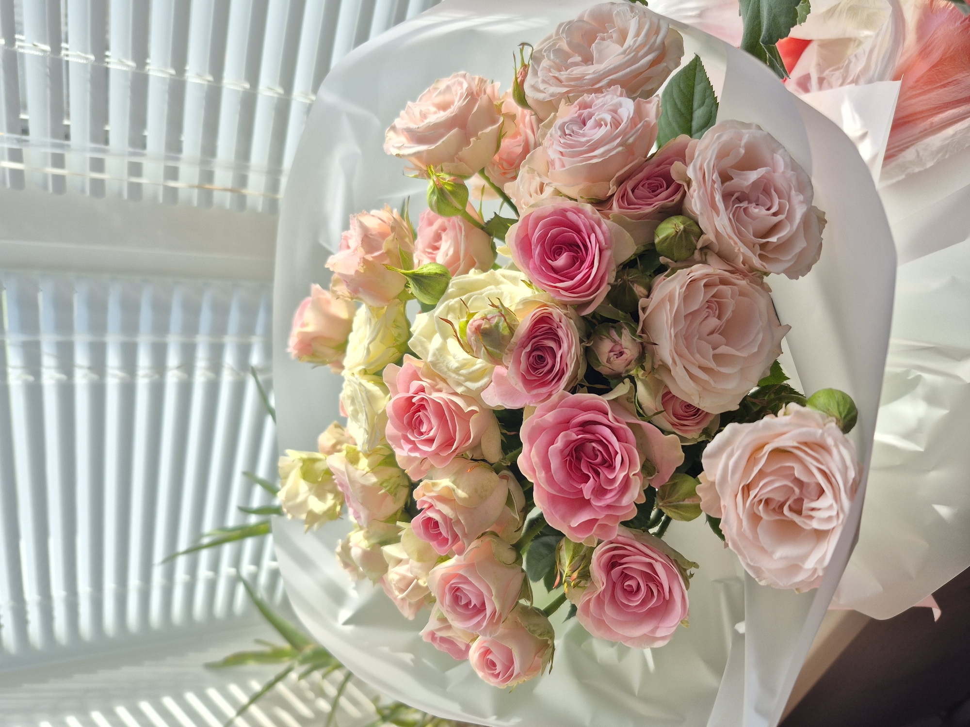 A bouquet of light pink and dark pink roses sits in a white wrapper. The bouquet is positioned near a window with white blinds, casting shadows across the scene. The location is unknown and there is no evidence to suggest the photo was taken in Votkinsk, Russia.