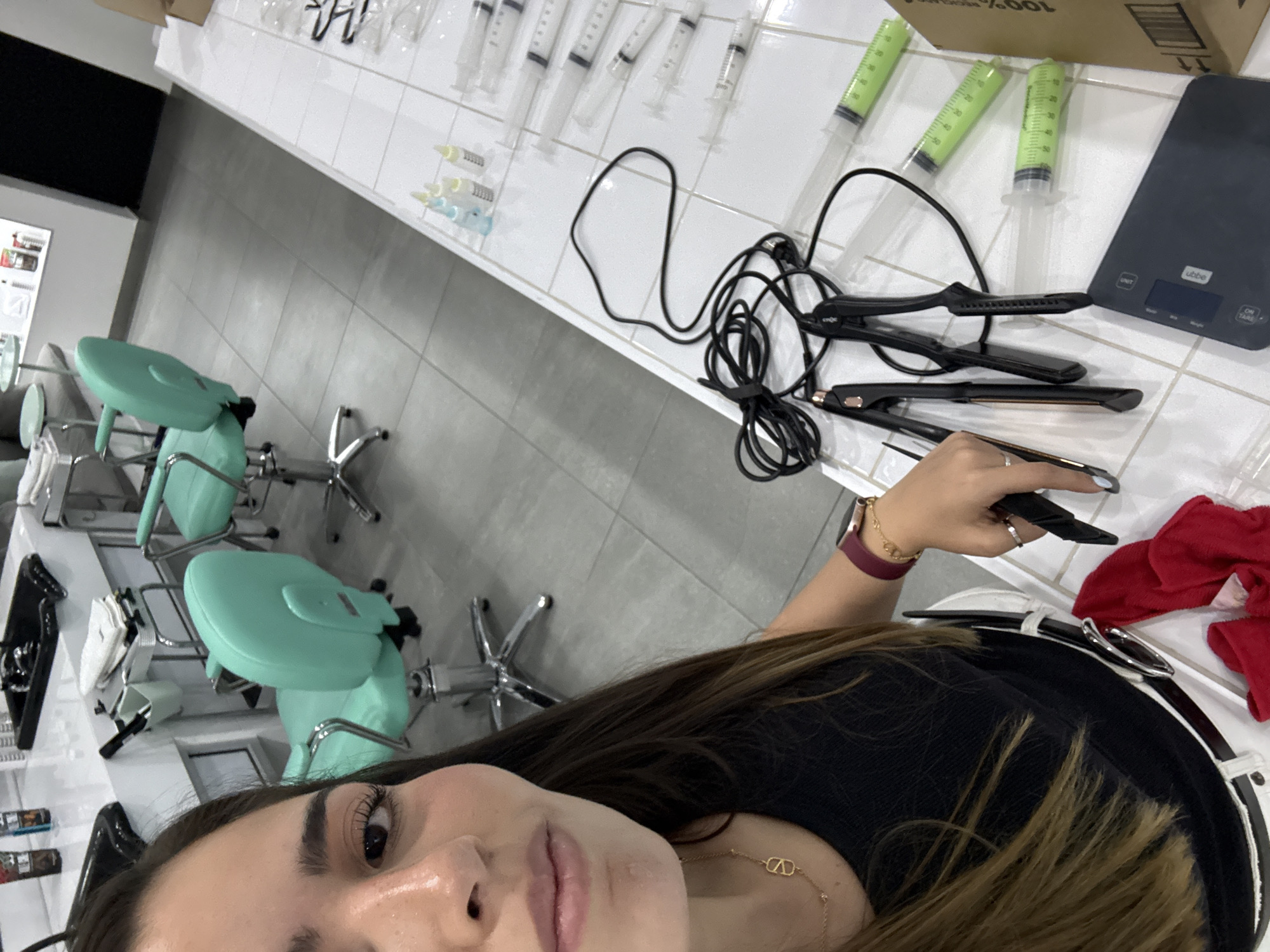 A selfie-style photograph taken indoors, likely in a salon or medical-aesthetic setting. A woman with brown hair, wearing a black top, a gold necklace, and a watch on her left wrist, dominates the foreground. The background features medical or cosmetic equipment, including syringes, styling tools (possibly curling irons), a digital scale, and salon chairs. The floor is tiled in light gray. The location is presumably in San Bernardino Tlaxcalancingo, Mexico.