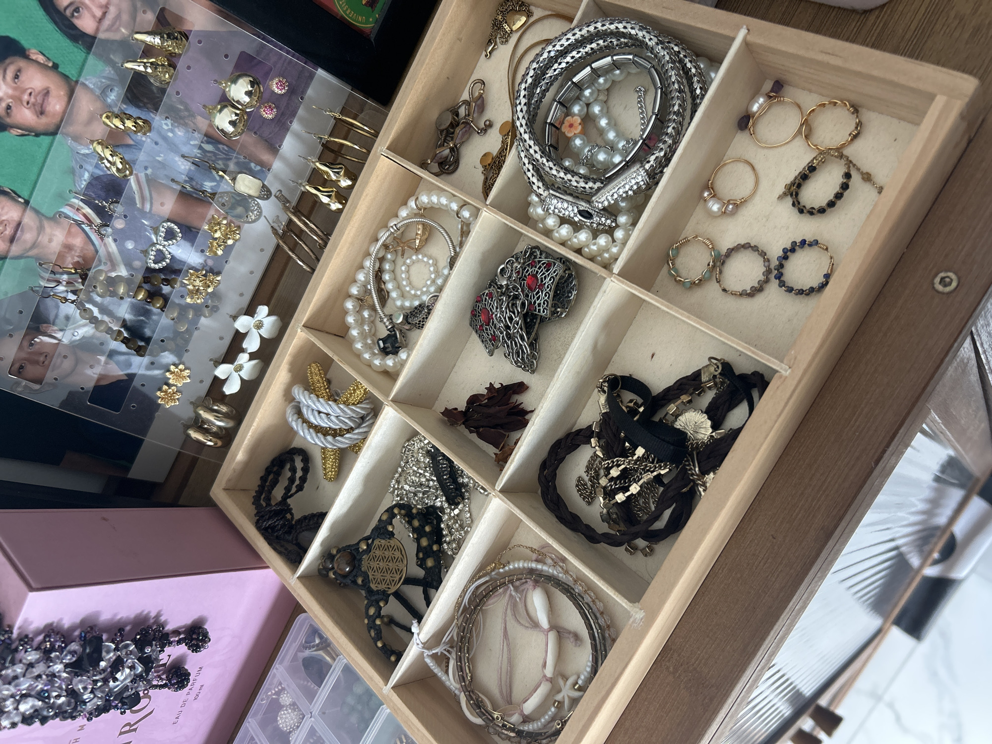 This is a high-angle, medium shot of a wooden jewelry box filled with various accessories. The jewelry box has several compartments containing necklaces, bracelets, and rings. In the background, there is a pink box with intricate beadwork and a clear plastic display case filled with earrings, behind which are images of people. The scene is set indoors, on a wooden surface. There are no people or animals present in the immediate scene. The image appears to be taken at eye level with the jewelry box.