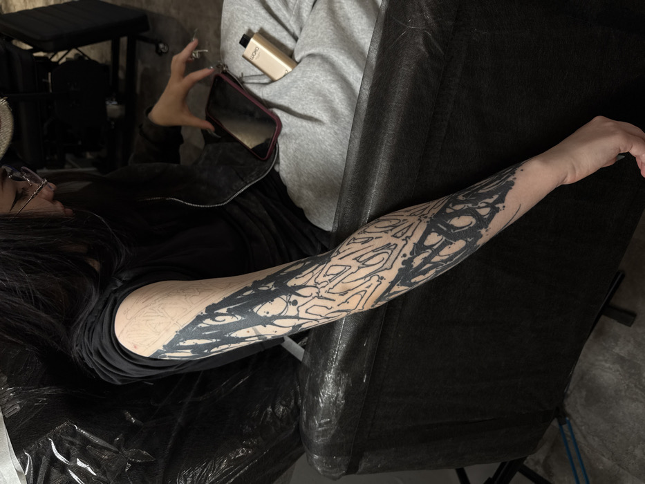 An indoor scene, located in Moscow, Russia, displays a female individual with long, dark hair and glasses. She is seated in a dark chair, which is partially covered by a black plastic sheet. Her left arm is extended, revealing a partially completed blackwork tattoo sleeve. The forearm is extensively covered in dense, abstract, graffiti-style black ink with drip patterns and outlined shapes, while the upper arm shows a stencil outline of the future design. She is dressed in a dark t-shirt, a dark zippered jacket, and light grey sweatpants. On her lap, a dark-cased smartphone is visible, alongside a light gold/beige 'LUXO' brand vape device. The flooring is dark, and the background is indistinct, consistent with a tattoo studio setting.