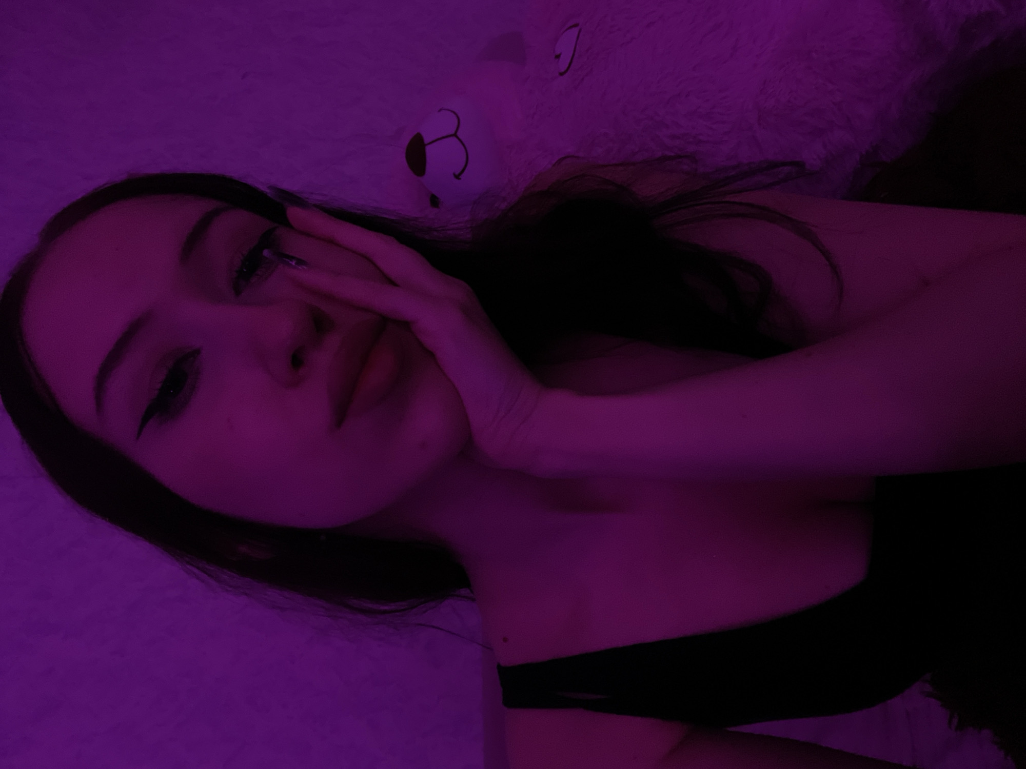 The image depicts a young woman with dark hair and eyeliner posing against a textured, purple-lit background. She is resting her right hand on her cheek, and her left arm is visible in a black top. To the right of the woman, a white, fluffy teddy bear with embroidered facial features is partially visible. The overall lighting is dim and cast in shades of purple. The location in the city of Votkinsk, Russia cannot be determined from the provided image.