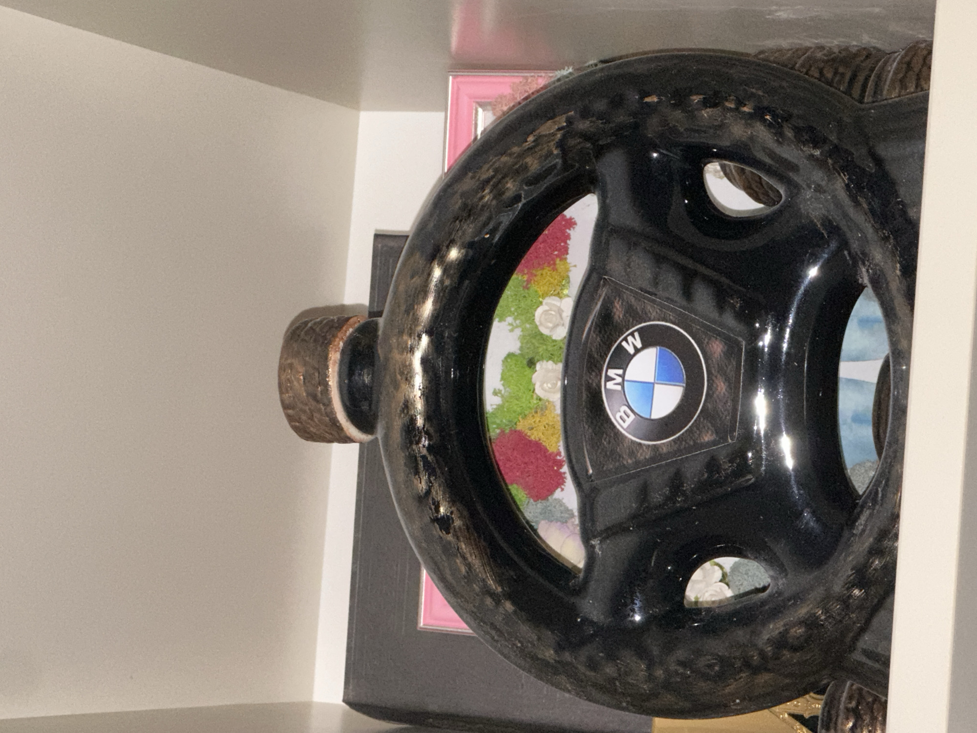 The image shows a black, circular object, possibly a stylized steering wheel, with a BMW logo at its center. The interior of the object contains an arrangement of colorful artificial flowers. The object is positioned against a background that includes a pink picture frame, a dark-colored frame, and sections of what appears to be a white shelf or cabinet. The photograph might have been taken in a domestic setting. The location, Harmanli, Bulgaria, is not discernible from the image.