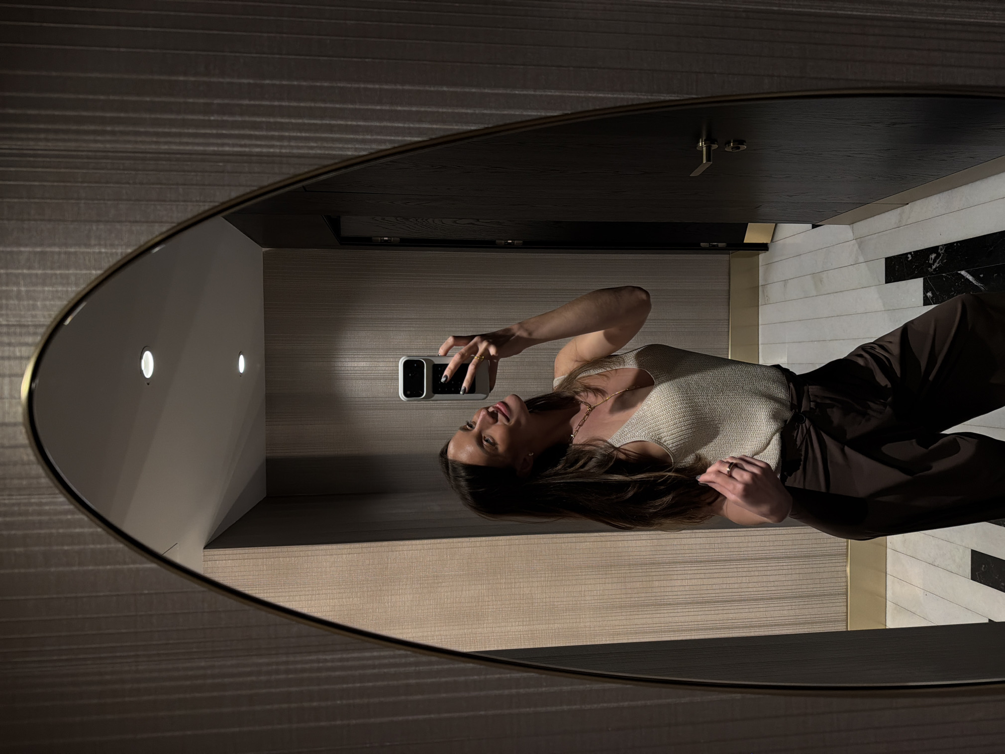 A woman is taking a selfie in a mirror in what appears to be a hotel bathroom. The mirror is oval-shaped with a bronze-colored frame and has two lights reflected in it. The woman is wearing a cream-colored top and dark brown pants. The bathroom has striped wallpaper, black countertops, and marble tile flooring. Based on the likely high-end aesthetic of the bathroom, and the availability of such luxury bathrooms in hotels, it is possible this is a high-end hotel in Budva, Montenegro.