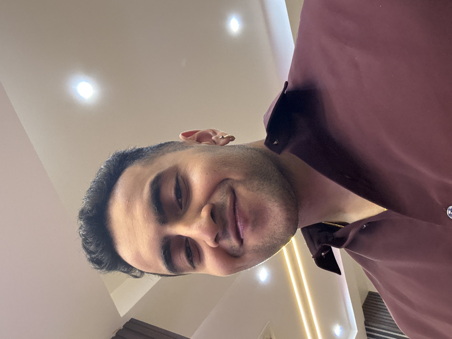 A man in a maroon collared shirt is looking slightly down at the camera with a subtle smile. His hair is dark and neatly styled, and he has a light beard. The background includes a white ceiling with multiple recessed lights. The camera is positioned low, looking upwards at the man. A small earring is visible in his ear.