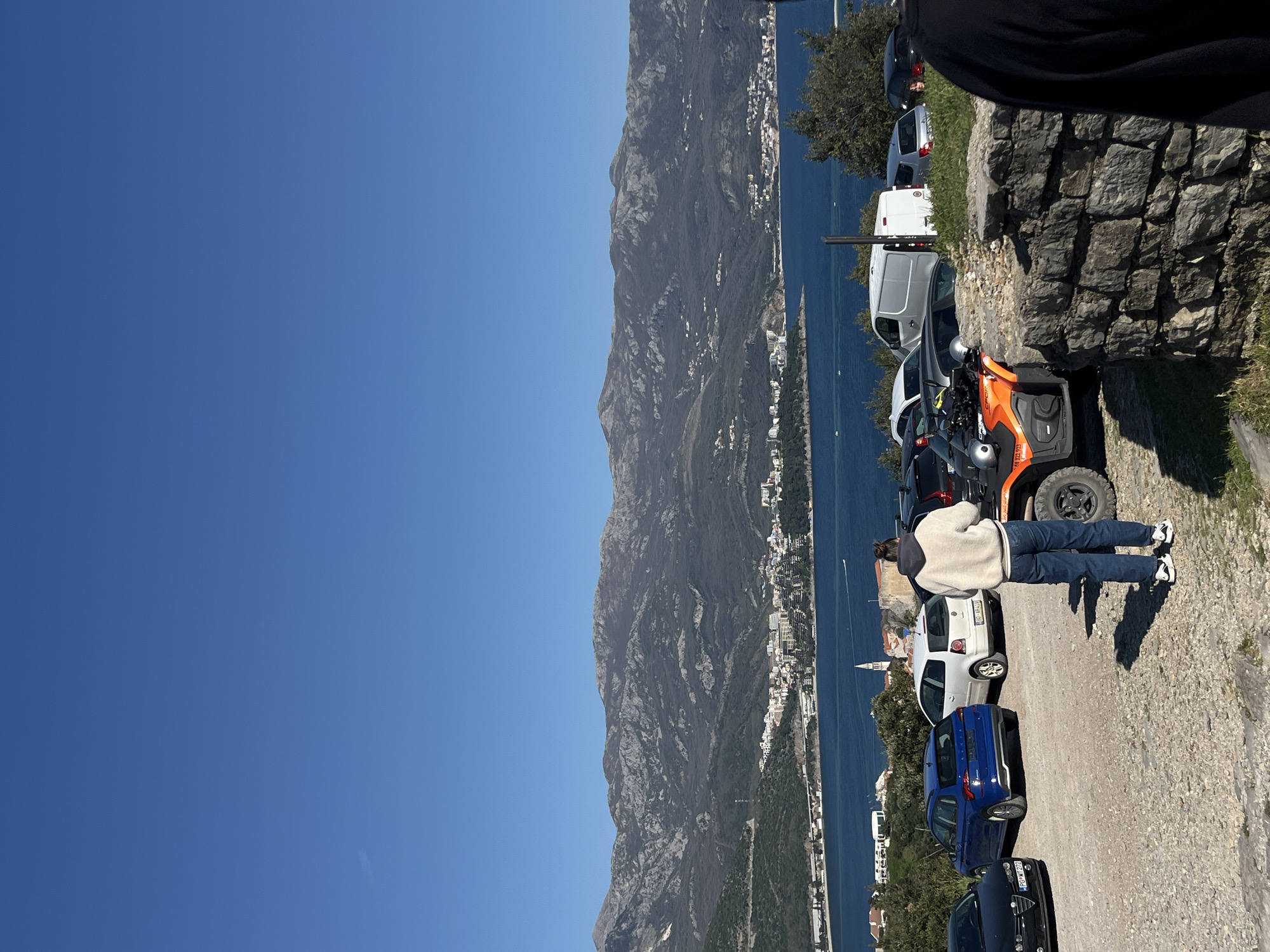 The image shows a mountainous coastal landscape on a bright, sunny day. The perspective is from a slightly elevated position, looking out towards a coastline dotted with buildings and extending into the distance. The sea is visible between the coastline and the mountains.
In the foreground, there's a narrow road or path lined with parked vehicles, including cars and an orange ATV. A person dressed in jeans and a light-colored jacket stands on the path, their back to the viewer. A stone wall is to the right, with foliage growing from it. The person is on the top of the road, looking to the scenery.
Based on the geography, buildings, and mountainous terrain, the location is likely near Budva, Montenegro.