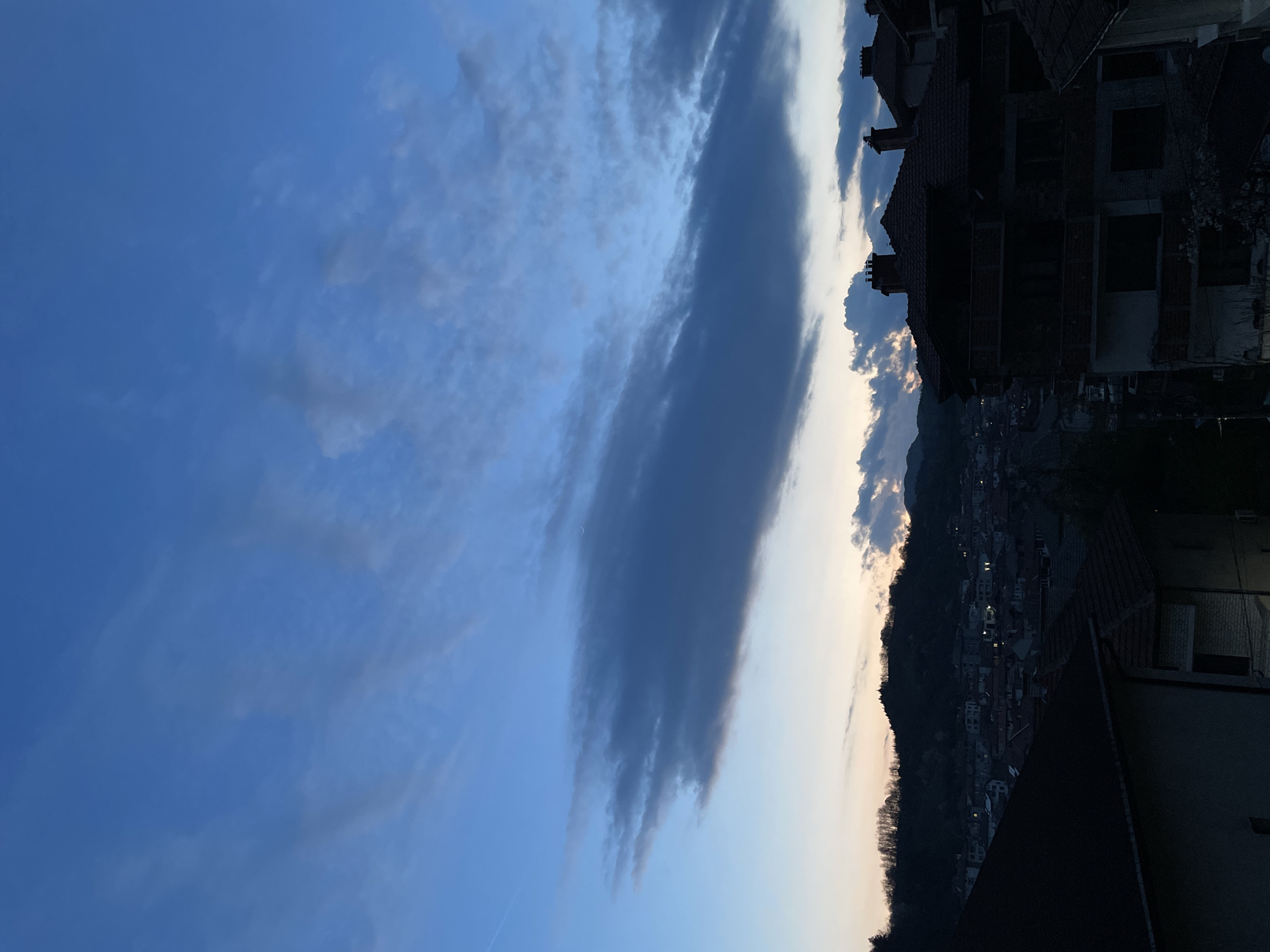 The image depicts an evening cityscape in Zlatograd, Bulgaria, viewed from an elevated position. The sky is predominantly blue with long, dark clouds running diagonally across the frame. In the background, a hill is silhouetted against the pale evening sky, with houses and buildings dotted across its slope, some with illuminated windows. The foreground features buildings with dark rooftops and window openings, their details obscured by shadow. The image is oriented sideways.