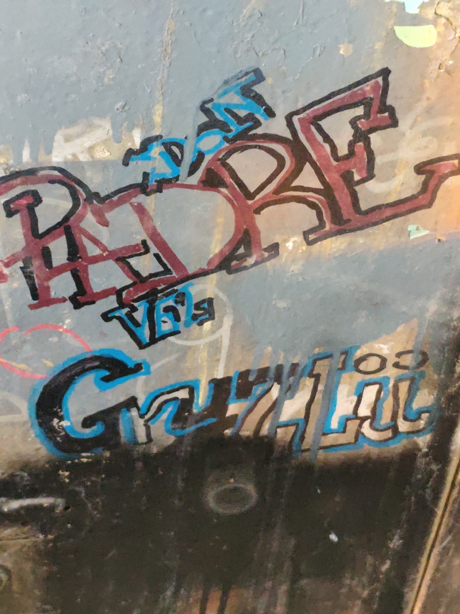 A close-up, high-angle, outdoor shot captures graffiti art spray-painted on a textured, weathered, light blue and grey metal surface. The graffiti features blocky, stylized lettering in red, black, and blue. In the upper section, red letters with black outlines form the word "HOPE," with some blue accents. Below this, a smaller tag in blue and black reads "VERY." The bottom portion of the image displays larger, bolder lettering in black with blue and light grey outlines, spelling out "GRIZZILL." The surface is grimy and shows signs of wear and tear, with streaks and discoloration. The background is out of focus, suggesting an urban environment.