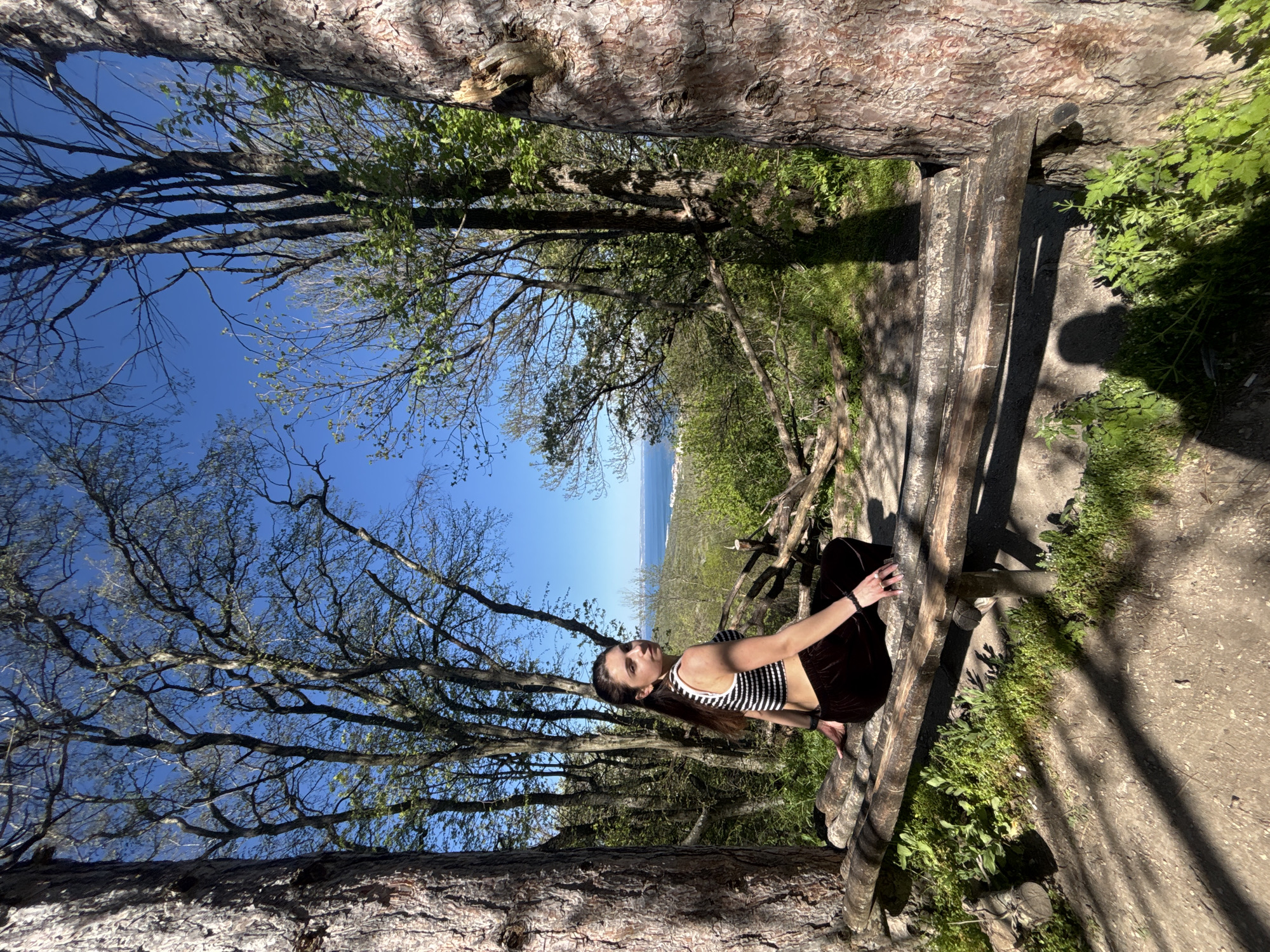 A woman is seated on a wooden structure within a forest area. She is wearing a black and white striped top and dark-colored pants. The background shows trees, a clear blue sky, and a body of water. This scene is likely located in the city of Varna, Bulgaria, as specified in the instructions. The exact location within Varna is not discernible from the provided image.