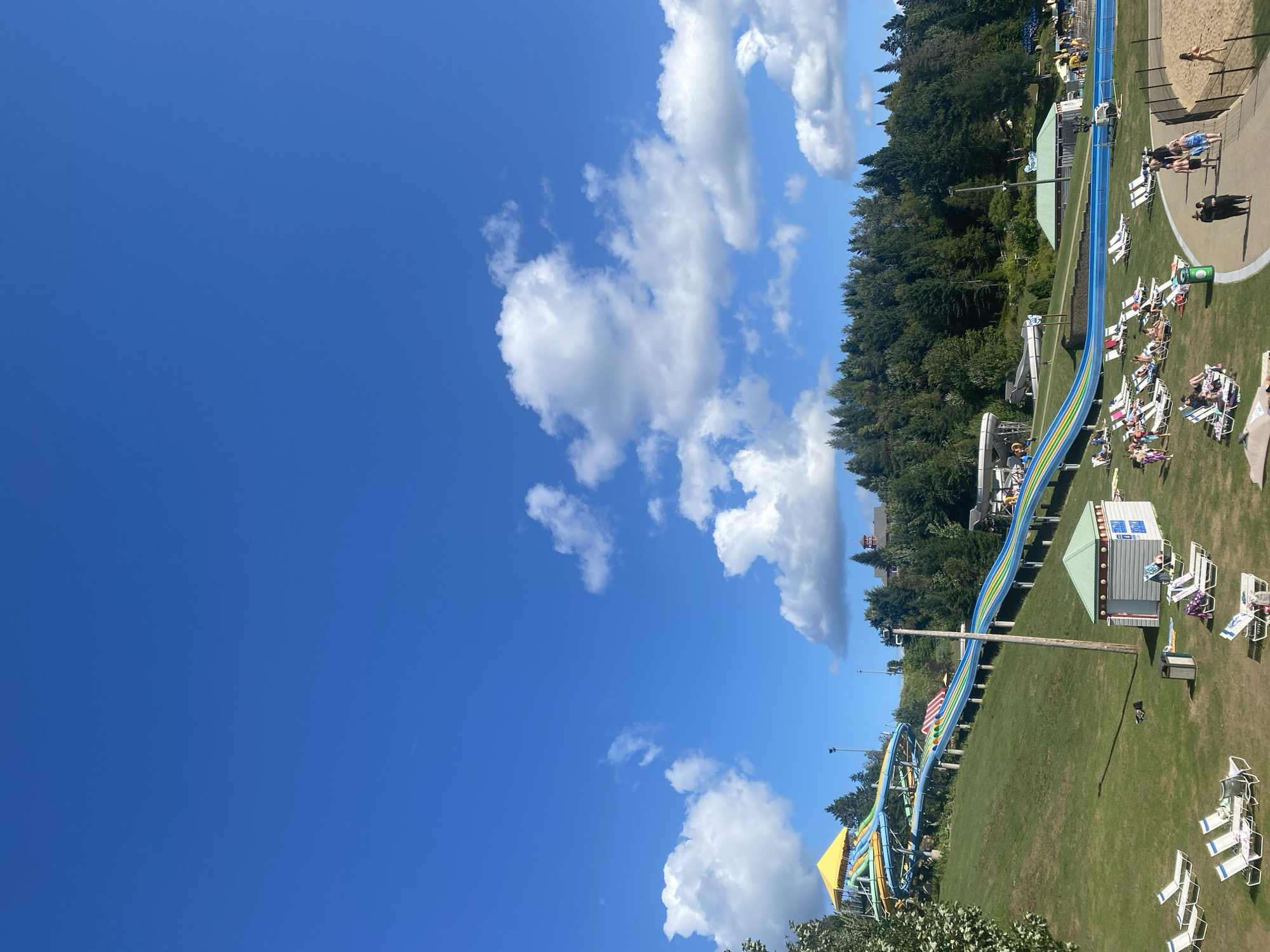 The image depicts a bright, sunny day at the Village Vacances Valcartier water park in Saint-Gabriel-de-Valcartier, Canada. The primary focus is a large, multi-colored water slide that snakes down a grassy hill. The slide features sections of blue, yellow, and green. At the top of the slide, there is a yellow covered platform. The surrounding area is a well-maintained green lawn dotted with numerous white lounge chairs, some occupied by people. A small, light-colored building with a green roof, likely a changing room or restroom facility, is visible in the foreground.
Further in the background, a dense forest of green trees forms a backdrop to the water park. The sky is a vibrant blue with scattered white, fluffy clouds. In the middle ground and to the right, a sandy area is visible, indicating a beach or play zone. Several people are walking along a paved pathway near the sandy area, and others are relaxing on lounge chairs on the grass. Some additional structures and slides are partially visible in the distance, suggesting the scale of the water park. The overall scene conveys a sense of leisure and outdoor recreation on a clear summer day.