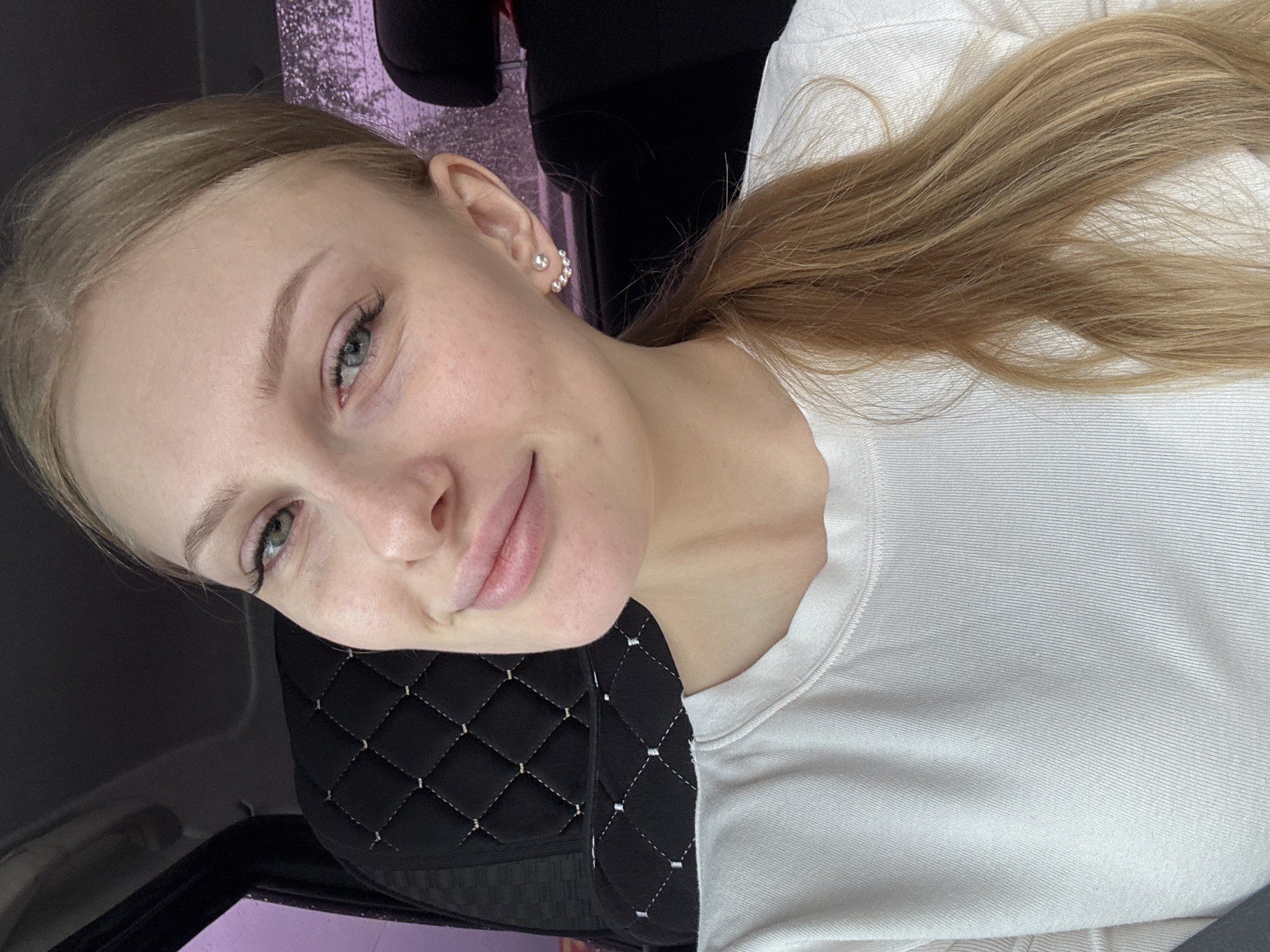 A young woman with light skin and blonde hair is shown from the chest up in an indoor setting. Her hair is pulled back in a ponytail. She is wearing a white long-sleeved shirt. She has a small earring on her left ear. Behind her is a black headrest with a diamond-stitched pattern. It is likely a car seat, and the location is Perm, Russia.