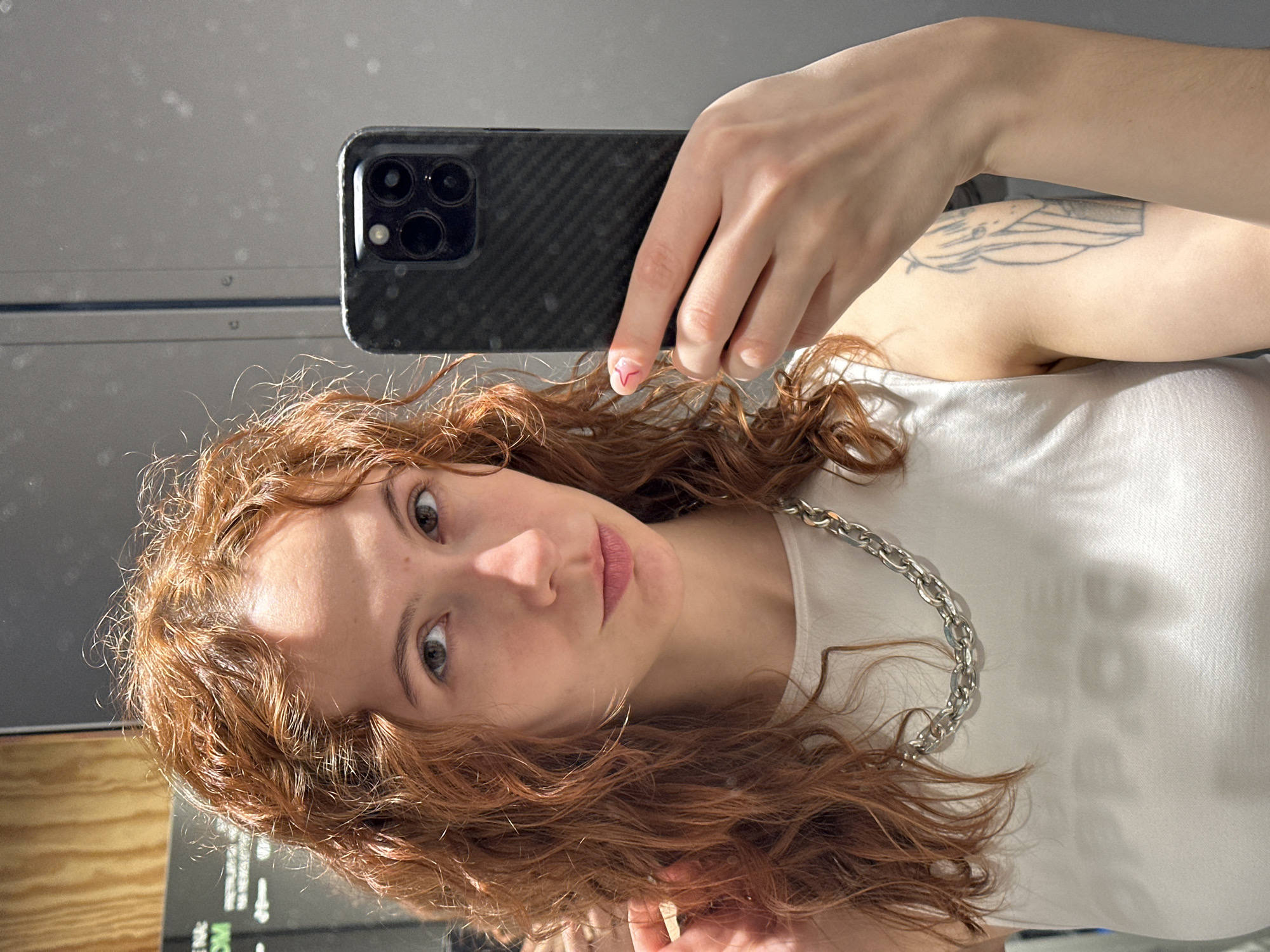 A young woman is taking a selfie in front of a mirror. She has long, wavy, reddish-brown hair and fair skin. She is wearing a white top and a silver chain necklace. She is holding a smartphone with a black case and multiple camera lenses. Her fingernails have a red design. A tattoo is visible on her upper arm. The background appears to be an interior space, perhaps a shop or office, with reflective surfaces and some visible text. While Novi Sad, Serbia, cannot be determined from the image alone, the overall setting seems urban and modern. The environment is well lit, emphasizing the woman and her immediate surroundings.