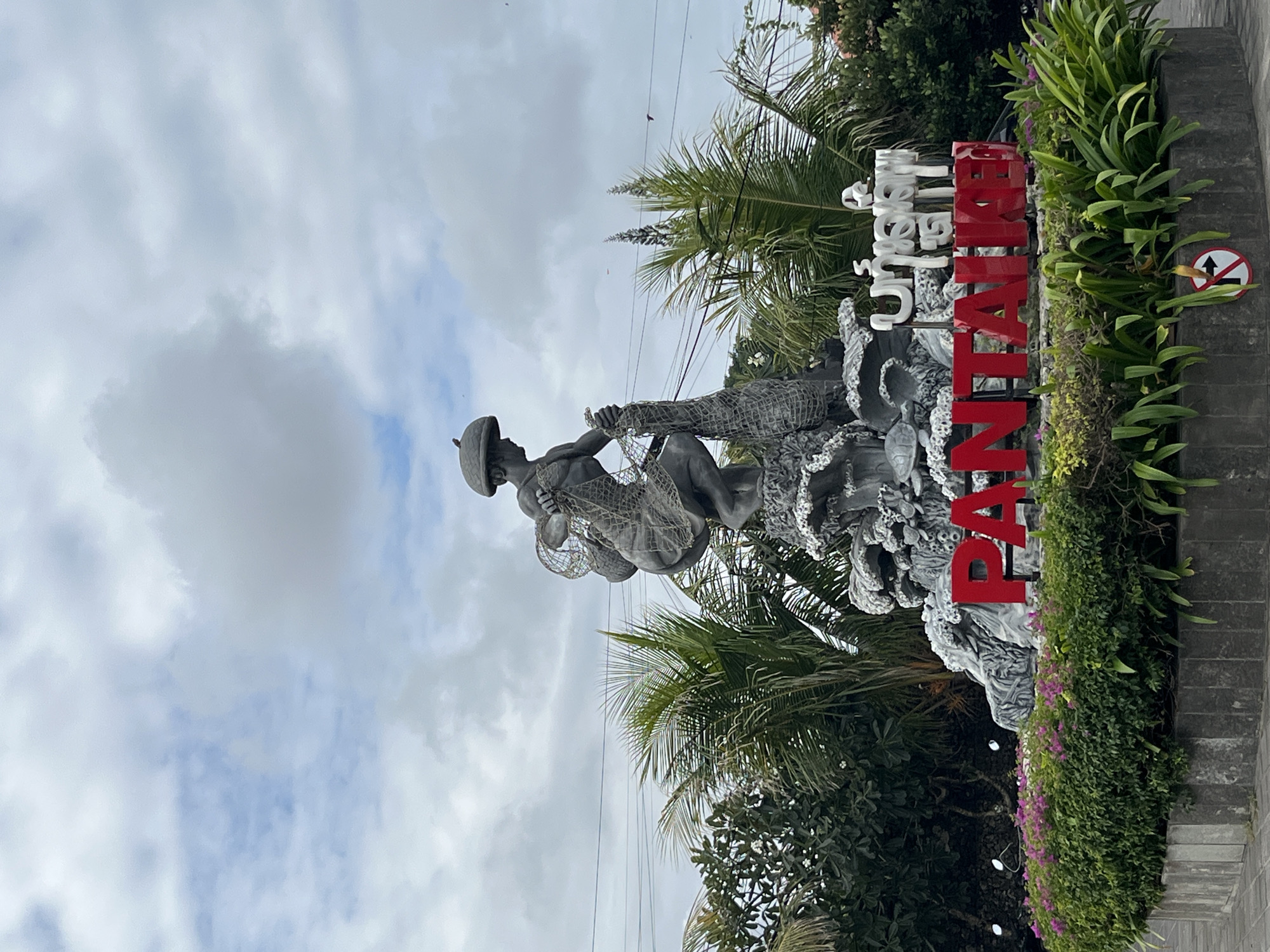 A medium shot captures the Kuta Bali Landmark, a prominent monument in Kuta, Indonesia, featuring a statue of a fisherman in traditional attire, complete with a net and a hat. The statue is perched atop a rock formation adorned with sculptures of marine life, including turtles. In front of the monument, large red letters spell out "PANTALAI," signifying "beach" in Indonesian. Above and behind the red lettering, white text in Balinese script is visible. The monument is surrounded by lush green foliage and vibrant pink flowers, with palm trees in the background. The sky above is overcast with scattered clouds.