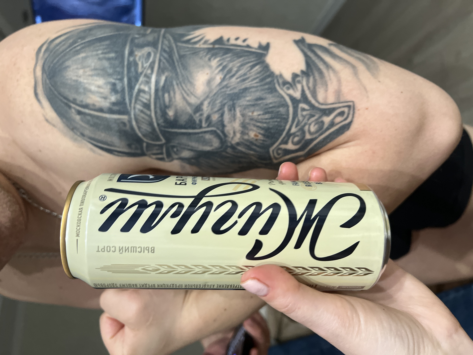 The image presents a close-up view of a person holding a can of beverage. The person's arm, visible across the upper half of the frame, is adorned with a prominent black and grey tattoo. The tattoo depicts a detailed warrior or Viking figure wearing a helmet, with elements resembling Mjolnir (Thor's hammer) featuring intricate knotwork designs.
Two hands hold a pale cream-colored can horizontally in the lower portion of the image. The can features large, dark, stylized Cyrillic script spelling "Жигули" (Zhiguli). Below this, smaller, inverted Cyrillic text reads "ВЫСШИЙ СОРТ" (Vysshiy Sort), translating to "Highest Grade." Along the left edge of the can, partially visible text in Cyrillic states "МОСКОВСКАЯ ПИВОВАРЕННАЯ..." (Moscow Brewing...). A warning label, also in inverted Cyrillic, reads "УПОТРЕБЛЕНИЕ АЛКОГОЛЬНОЙ ПРОДУКЦИИ ВРЕДИТ ВАШЕМУ ЗДОРОВЬЮ" (Consumption of alcoholic products is harmful to your health), confirming the beverage is alcoholic.
The background is largely blurred, suggesting an indoor setting, with indistinct shapes and colors. A faint silver chain is visible around the neck of the person. The lighting appears even, but the time of day and specific location are not discernible. No particular activity beyond holding the can is depicted, and no emotions are conveyed.