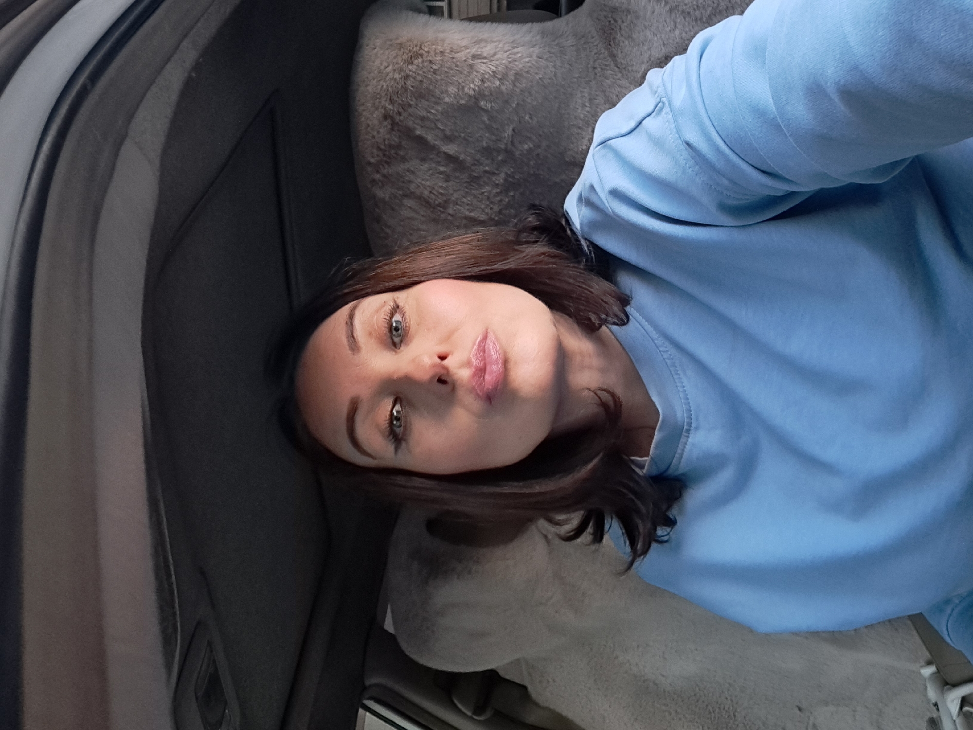 A woman with shoulder-length brown hair and fair skin is sitting in the driver's seat of a car. She is wearing a light blue sweatshirt and has makeup on, including eyeliner and blush. Her lips are pursed as if she is about to kiss. The interior of the car is visible, with fluffy grey seat covers and the car's ceiling. The image appears to be taken from a selfie perspective. The scene does not provide any information to determine the location in the city of Irkutsk, Russia.