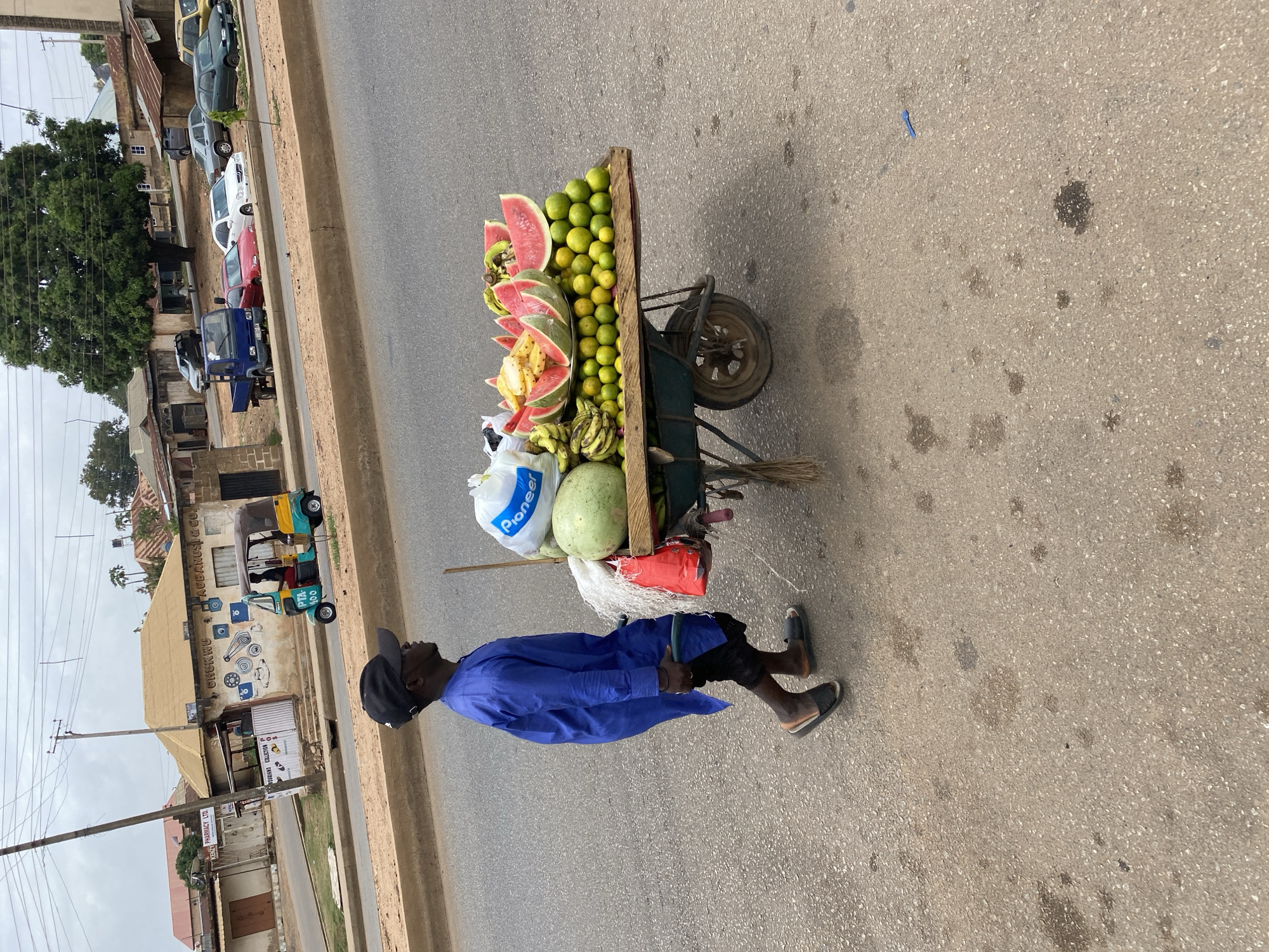 A man in a blue robe and black cap pulls a wooden cart filled with fruits along the roadside in Bukuru, Nigeria. The cart contains cut watermelons, limes, bananas, and a watermelon. In the background, there are parked cars, buildings, and a tricycle.
