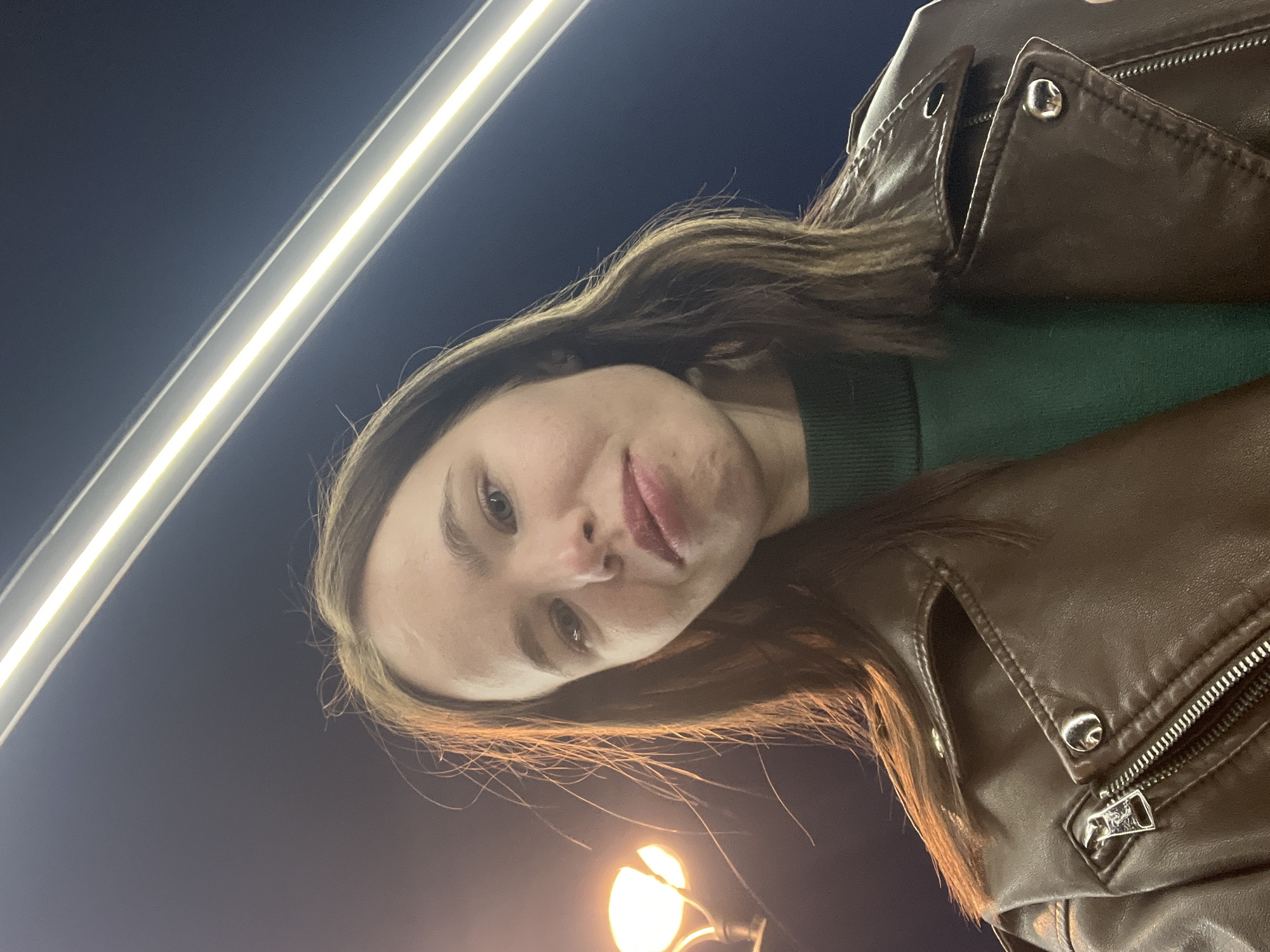 This is a rotated close-up shot of a young adult, likely female, taken in Chaikovsky, Russia, at night. The subject's face occupies the central-left portion of the frame, angled slightly upwards towards the camera. She has long, dark brown hair with lighter undertones, which frames her face and falls over her shoulders. Her expression is neutral, with her lips slightly parted, and she looks directly into the lens. Her eyes are a light color.
She is wearing a dark green, ribbed crew-neck sweater or top, visible beneath a brown leather-look jacket. The jacket features prominent details such as a zippered pocket, a main zipper closure, and several metal snap buttons or studs, indicative of a biker or casual style.
The background is dark blue, suggesting a night sky. Two distinct artificial light sources illuminate the scene. In the upper left, a long, bright, linear light source, possibly a fluorescent tube or LED strip, extends diagonally across the frame. In the lower right, a round, glowing street light fixture is visible, casting a warm yellow light. The combination of the dark background and artificial lights confirms the nighttime setting. There is no visible text in the image. The photo appears to be a selfie or a close-up portrait taken outdoors in an urban environment.