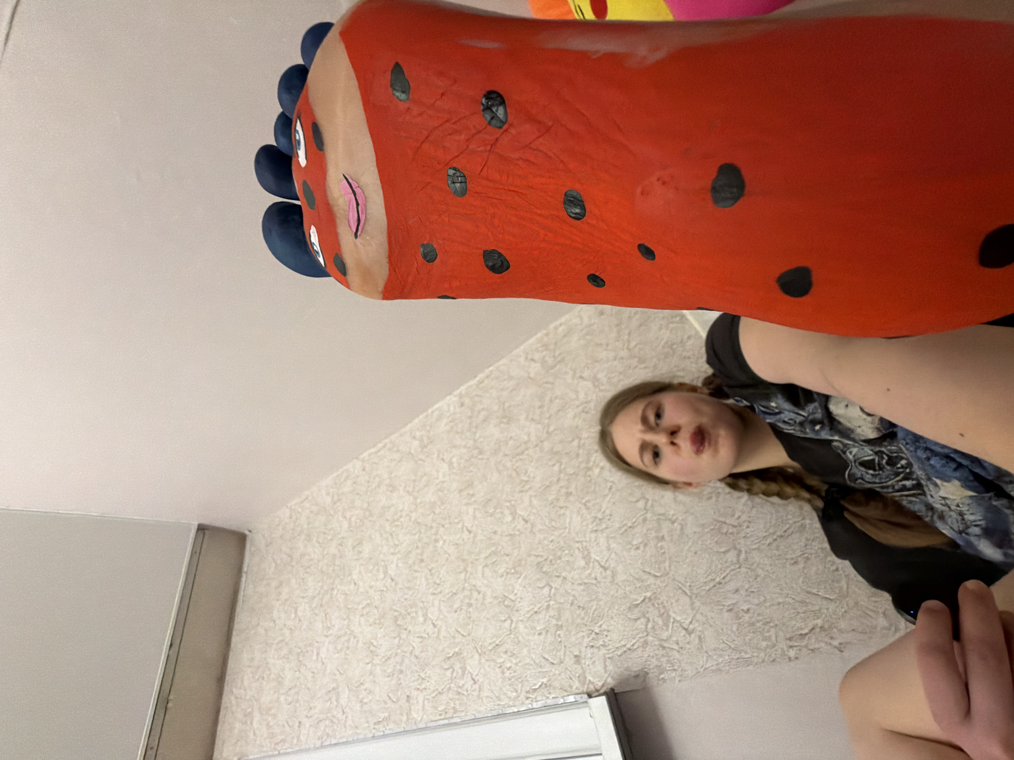 A low-angle shot taken indoors shows a person holding up their leg, which has been painted to resemble a ladybug. The foot is painted red with black spots, and the toes are painted blue. The person is lying down, and their face is visible to the right, with blonde hair braided down their back. They are wearing a black t-shirt with a blue and white patterned scarf. The background includes a textured wall and a white object that appears to be a window or shelf. The location is not provided, and information about the city of Perm, Russia, is not present in the image.