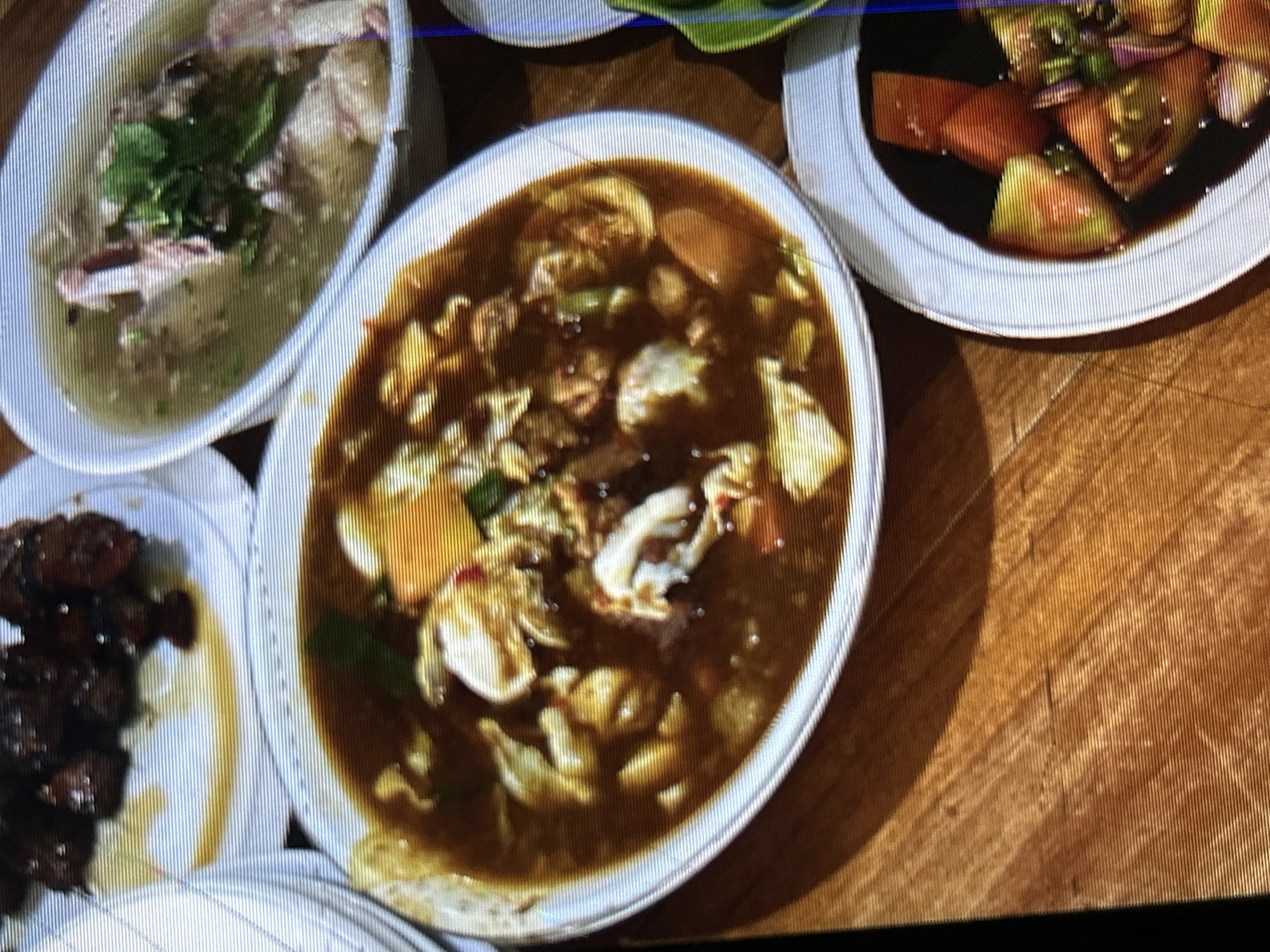 The image displays a close-up, overhead view of a wooden table set with several dishes of food, suggesting a meal in Depok, Indonesia. In the foreground, a white oval bowl is filled with a rich, dark broth containing chunks of meat, what appears to be scrambled egg, and possibly vegetables. To the right, another white bowl contains chopped tomatoes and other garnishes in a dark sauce. In the background, a white plate holds dark, bite-sized pieces of meat, and a separate white oval bowl contains a light-colored broth with pieces of meat and leafy green herbs. The lighting is dim, and the image has a grainy texture.