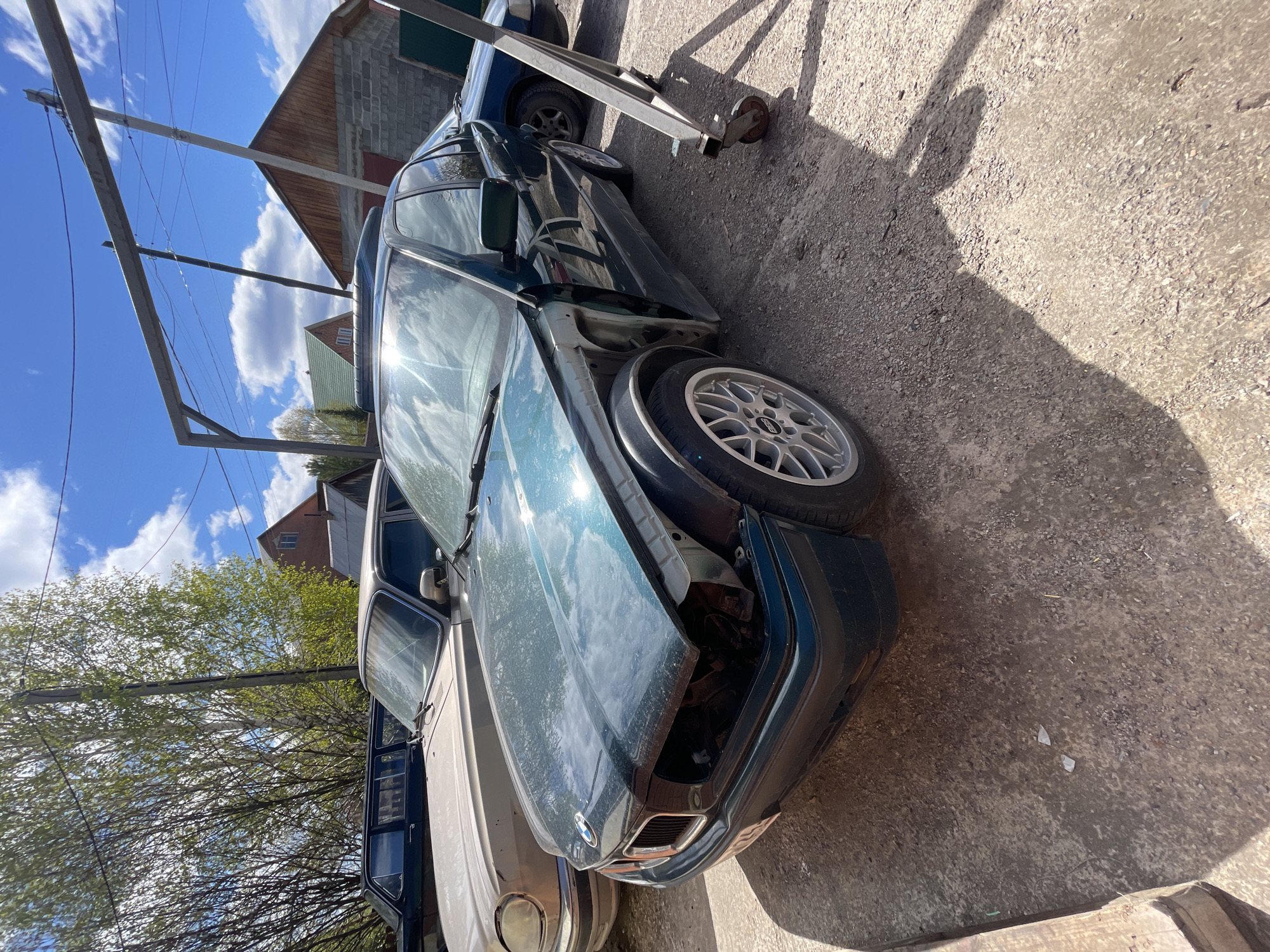 The image depicts a damaged dark green BMW sedan parked on a concrete surface outdoors. The front end of the car is heavily damaged, with the front bumper and panels missing. The driver's side door is open. The car has a wheel with a silver multi-spoke design. A tan-colored car is partially visible beside the BMW. Overhead, there are utility poles and wires against a background of a blue sky with clouds and nearby buildings. Based on the location provided, the scene is situated in Минино, Russia. There are no people visible in the frame.