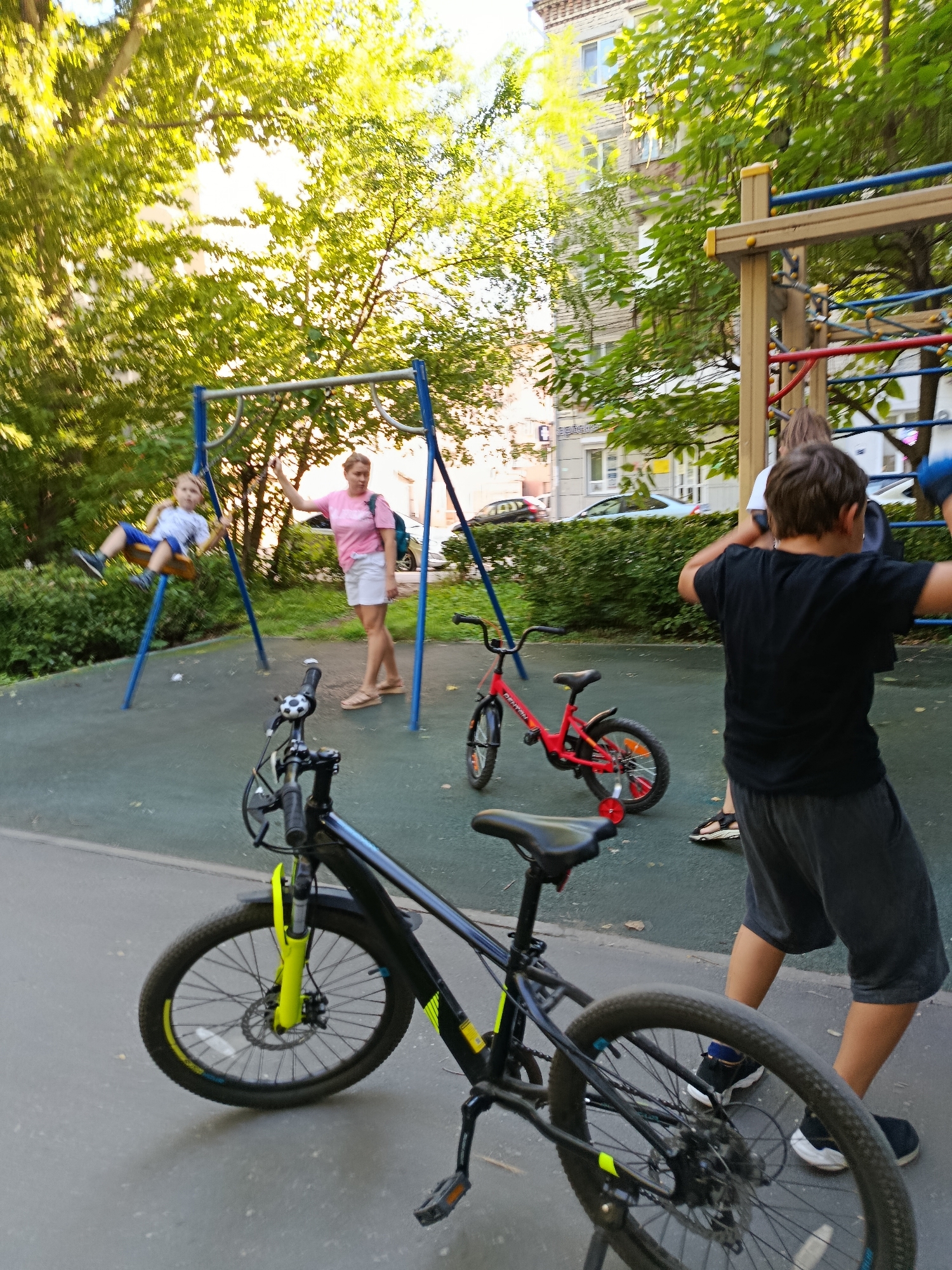 A group of people, including at least two children and one adult woman, are at a playground in Saratov, Russia. One child is on a swing, another child is standing near a red bicycle, and an adult woman is standing by the swing set. There are two bicycles in the foreground, one black and yellow, and the other is red. The playground has a climbing structure in the background, and the scene is framed by trees and apartment buildings. The ground is paved with a soft, dark material.