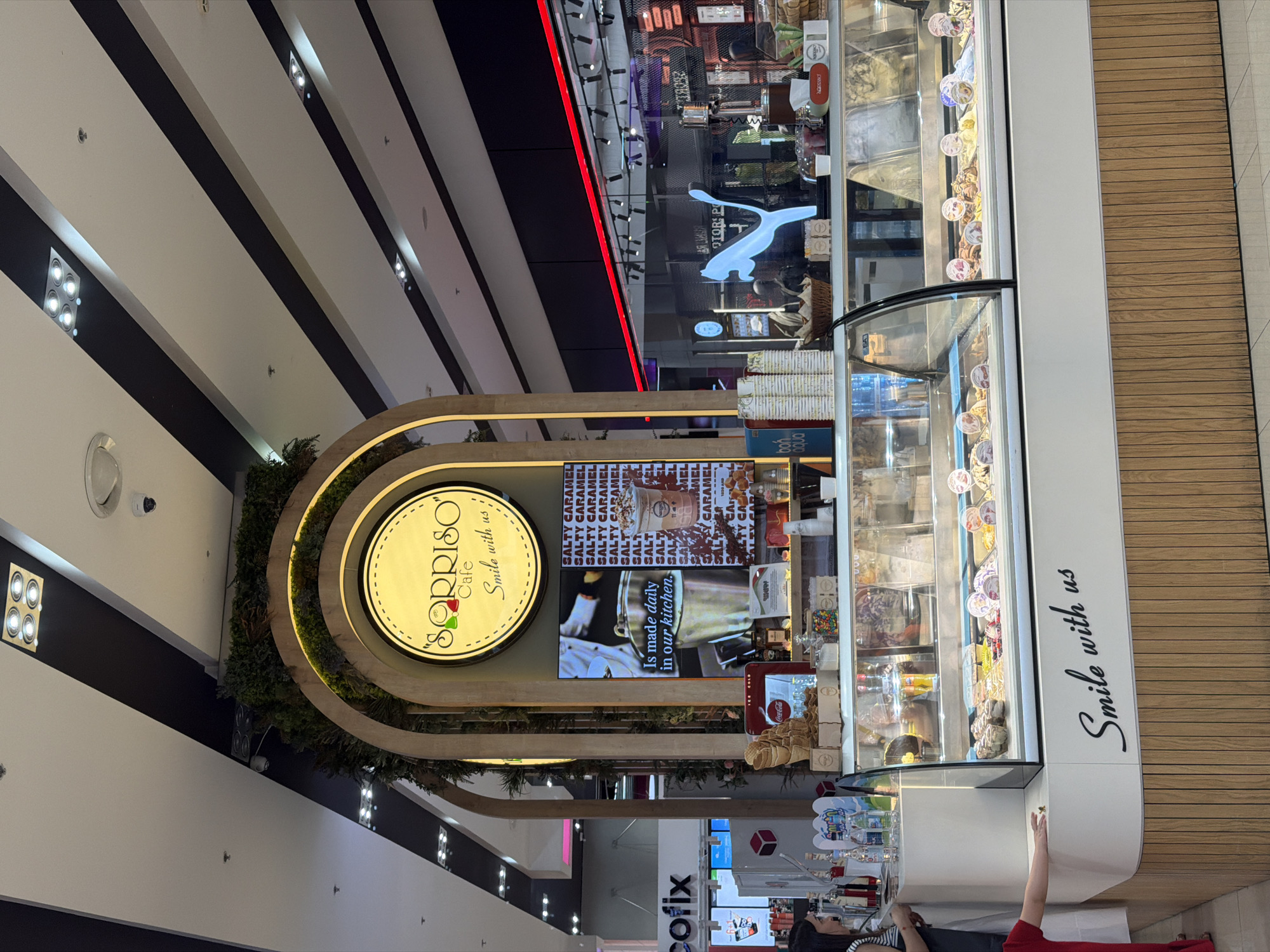 The image depicts a brightly lit interior of a modern cafe or ice cream parlor, likely situated within a shopping mall. The overall aesthetic is clean and contemporary, with a focus on natural materials and vibrant branding. A prominent, arched wooden structure frames the cafe's signage, which features the name "SORRISO" in a stylized font, accompanied by the tagline "Smile with us."
Beneath the signage, a digital display shows a video or image related to their products, possibly showcasing the making of ice cream. The cafe's counter is laden with an array of ice cream flavors displayed in chilled glass cases. Waffle cones are stacked neatly in a dispenser, and various toppings and condiments are organized on shelves. To the left, a woman wearing a striped top is partially visible, interacting with the display. A child in a red outfit is reaching towards the counter.
In the background, other retail spaces are visible, suggesting a commercial setting. A blue neon sign in the shape of a leaping feline, possibly a logo for a sports brand, is illuminated in the distance. The ceiling features an intricate design of white and black diagonal stripes, interspersed with modern lighting fixtures. The floor is tiled with a light-colored pattern. The cafe's facade is adorned with the phrase "Smile with us" in elegant script. This scene is consistent with the typical setup of an establishment found in the Yerevan Mall in Yerevan, Armenia, given the presence of well-known international brands and the mall's architectural style.