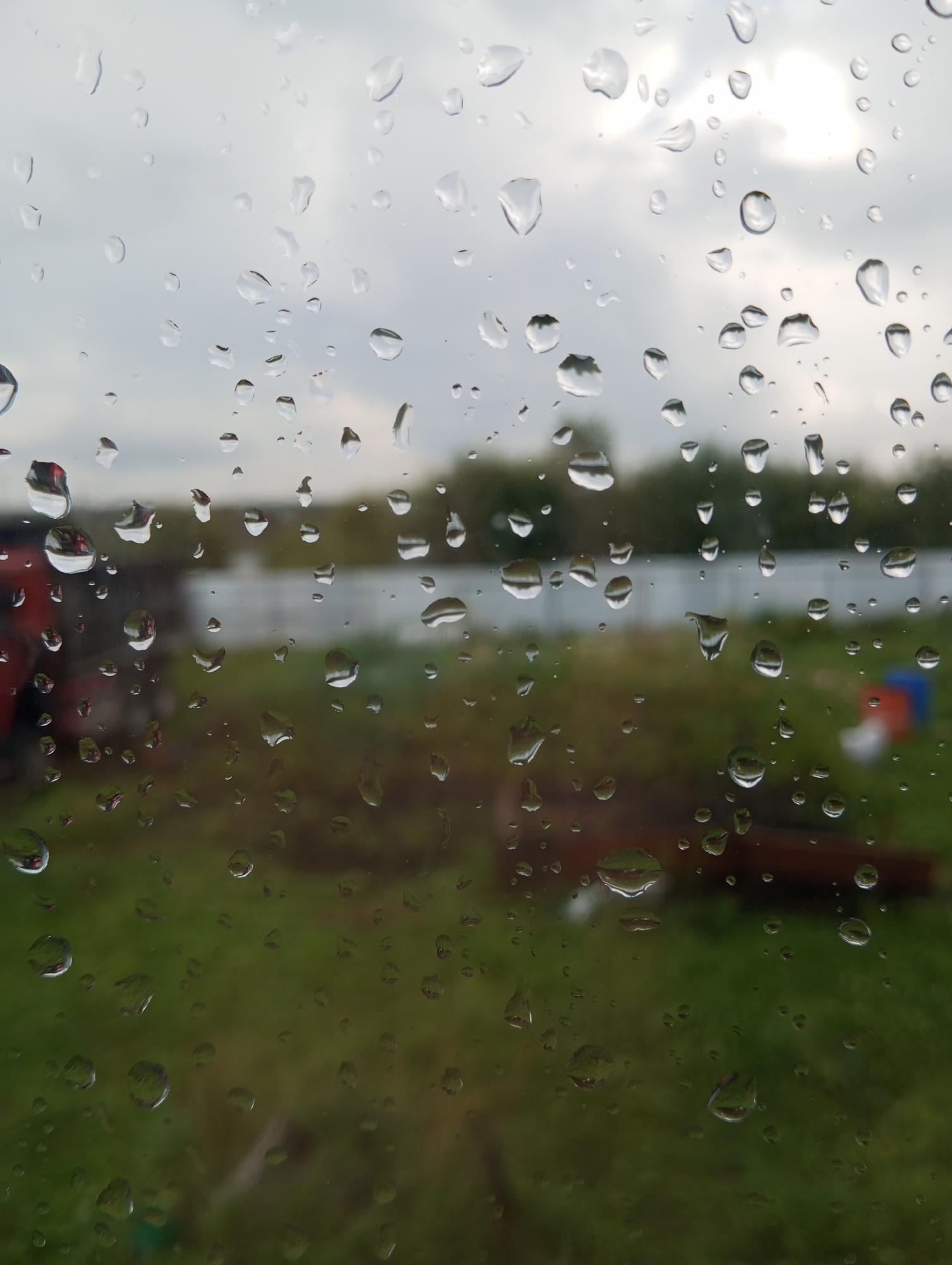 The image shows a view through a window covered in raindrops, looking out onto a rural or suburban landscape. The foreground is dominated by the water droplets, which distort the view of the background. The background is blurred but suggests green grass, trees, and some structures. The sky appears overcast with hints of sunlight breaking through. There are no people or animals visible in the image. The location is stated to be in Verkhnyaya Pyshma, Russia.