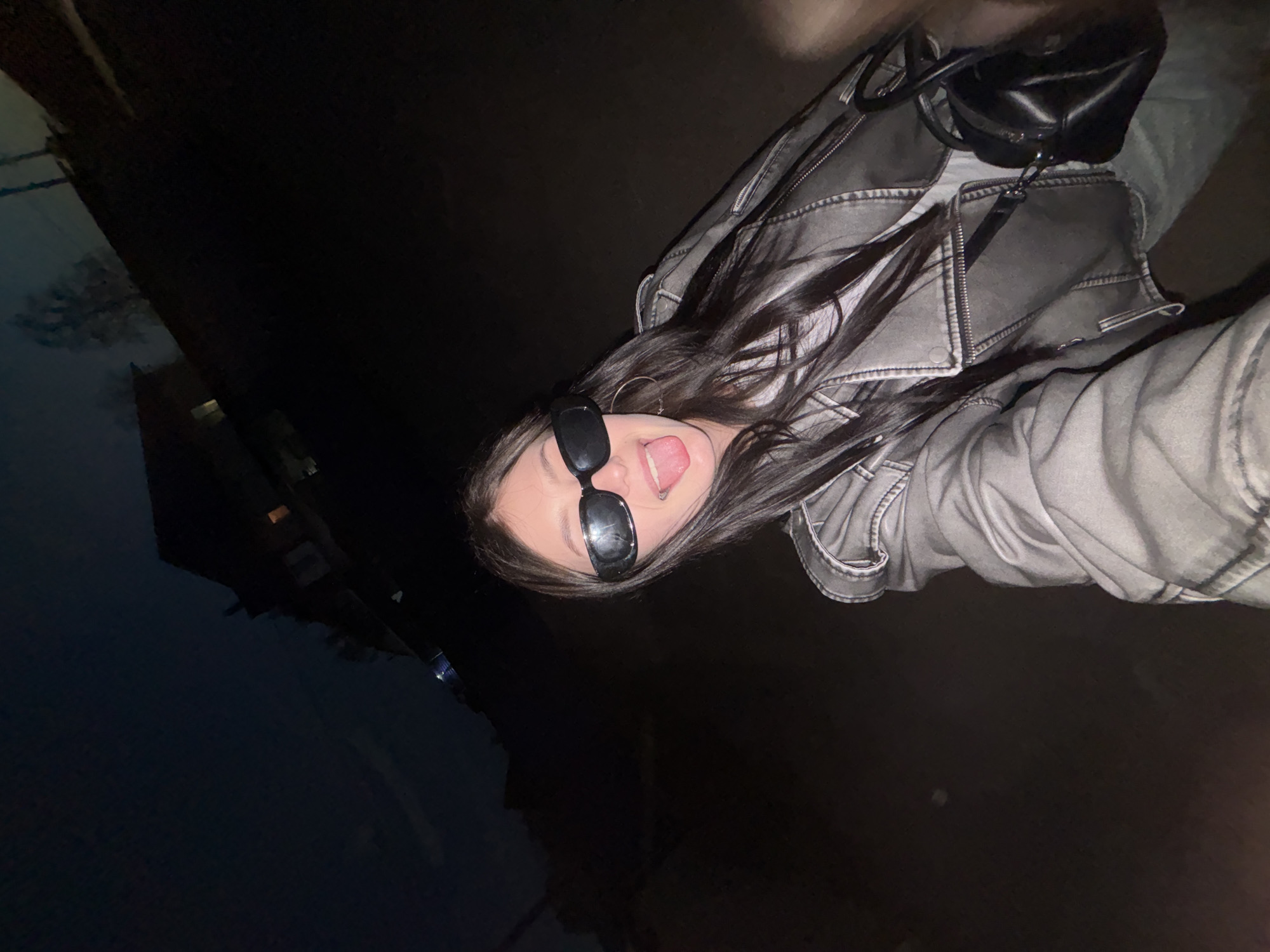 A selfie taken at night shows a woman wearing sunglasses, a gray jacket, and dangling earrings, sticking out her tongue. A black purse hangs from her jacket. The background is dark, with faint outlines of buildings visible in the upper left. It is not possible to confirm the location as being in Фоки, Russia from the image.