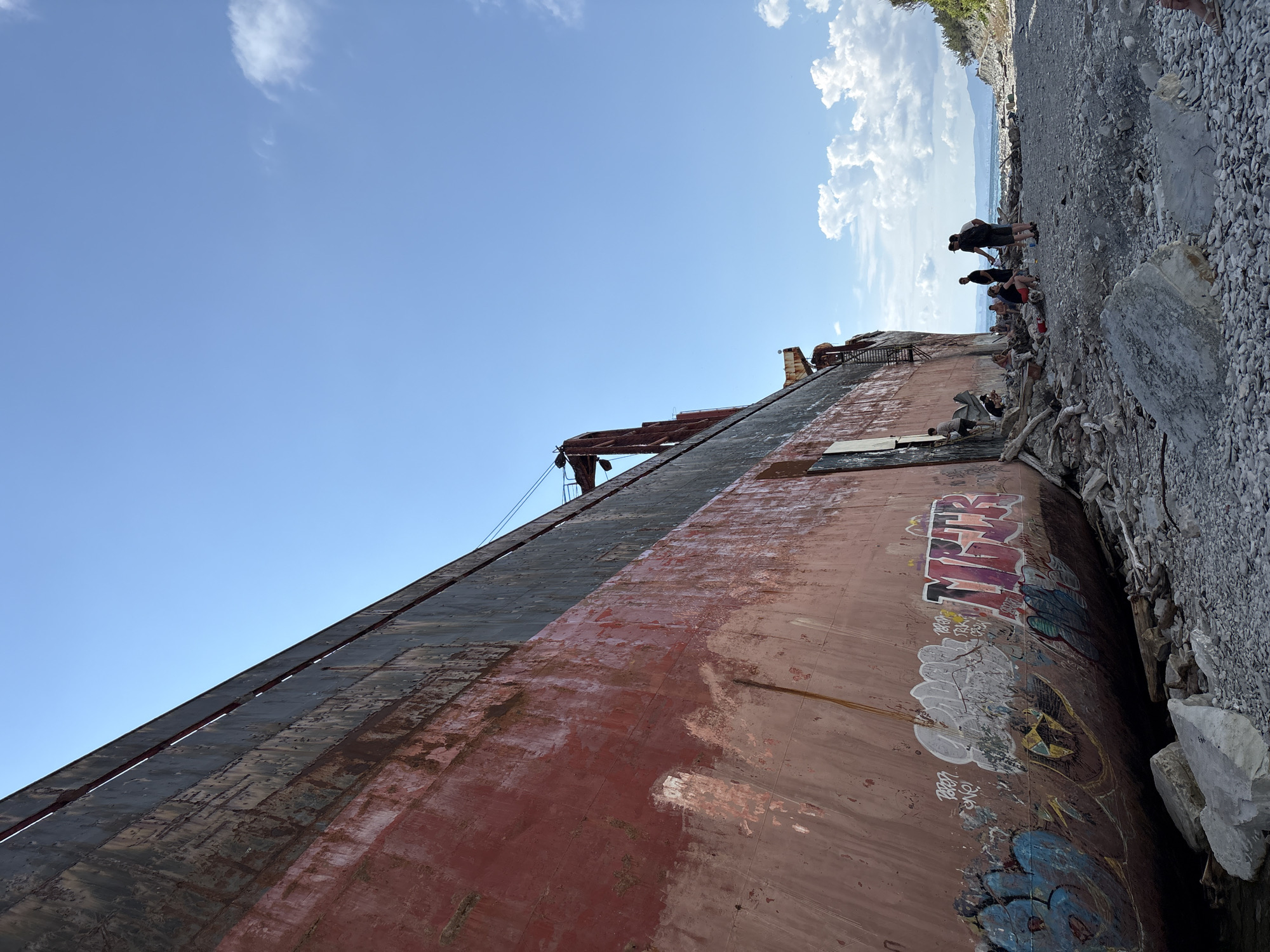 A large, rusting, red and black cargo ship lies beached on a rocky shore. The ship's hull is covered in graffiti. A group of approximately ten people are scattered around the ship, some exploring its exterior, others gathered in conversation on the beach. The environment is a coastal area with a rocky beach, dry driftwood, and a body of water visible in the distance. The sky is blue with scattered white clouds. This scene is located in Kabardinka, Russia, a known site for shipwrecks.