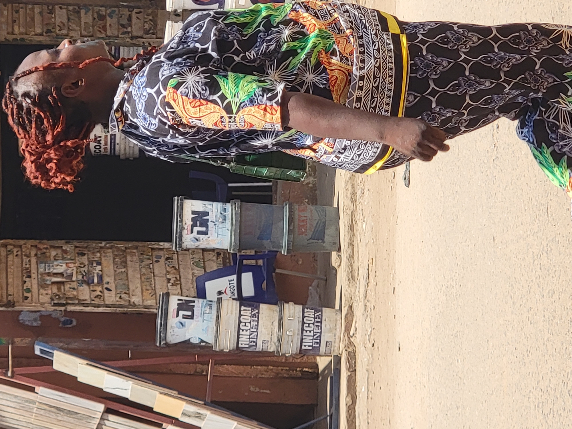 This image, set in Maiburiji, Nigeria, captures an outdoor scene featuring an individual amidst building materials. The central subject is a person, likely an adult female, standing with her head tilted upwards, looking towards the top left of the frame. She has dark skin and distinct reddish-brown dreadlocks or braids styled upwards.
She is dressed in a vibrant, full-body garment (possibly a co-ord set or jumpsuit) with a complex pattern. The attire features a black background adorned with intricate white, blue, green, orange, and yellow motifs, characteristic of African wax print fabrics. A striking yellow and black striped band runs vertically along her side.
The setting appears to be an open-air commercial area or a shop's exterior. The ground is an unpaved, dusty light brown. In the background and mid-ground, various items are visible. Several white and grey paint buckets are stacked, some with visible branding. Clearly readable text on these buckets includes "FINECOAT FINETEX." Partial text like "NANGOTE" and "COAT" is also discernable on other containers. Blue plastic chairs or stools are scattered between the stacks of buckets. In the foreground and background, various display tiles or wooden samples are arranged on racks.
The bright, even lighting suggests it is daytime, likely sunny, given the clear illumination. The person's expression is neutral as she looks up, and no specific activity beyond standing and observing is apparent. The overall environment indicates a retail point for construction or home improvement supplies in an urban or semi-urban African context.