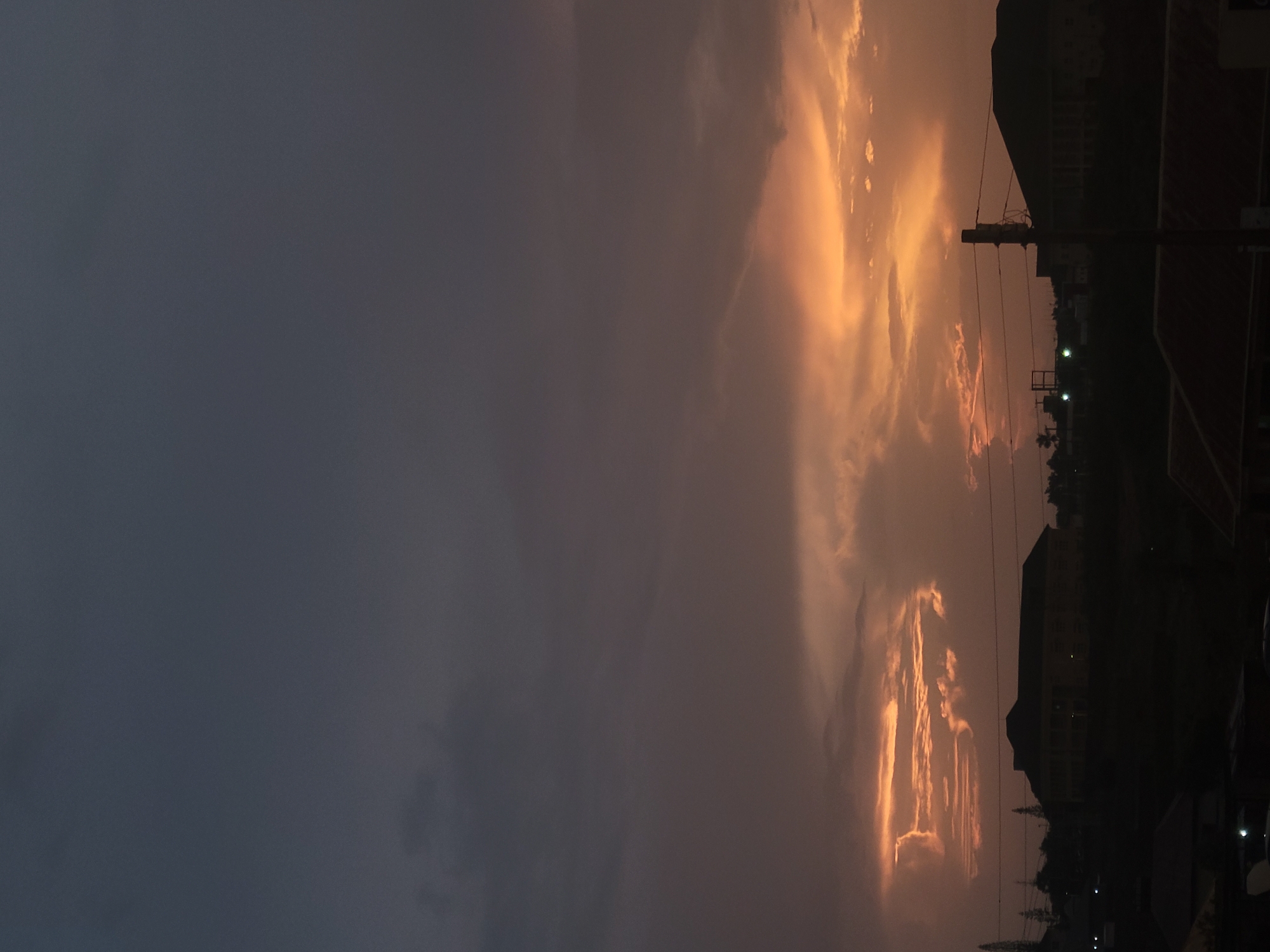 The image captures a dramatic sunset over a cityscape, likely Jos, Nigeria, given the context provided. The sky is a canvas of deep grays and blues at the top, transitioning into warm hues of orange and yellow where the sun is setting, illuminating the undersides of the clouds. A prominent band of bright orange light cuts across the middle of the frame, creating a striking contrast with the darker sky above and the silhouetted landscape below.
In the foreground, the silhouettes of various buildings are visible against the horizon. These include what appears to be a large, multi-story structure with a dark roof and numerous windows, situated on a raised area. To the left of this building, the dark outlines of trees and smaller structures are discernible. To the right, a utility pole with wires is a distinct vertical element in the scene, further emphasizing the urban environment. The ground level is largely obscured by shadow, with hints of rooftops and possibly roads. The overall impression is one of a vast, dramatic sky dominating the scene, with the city elements serving as a dark, grounding contrast. There are no visible people or active events depicted.
