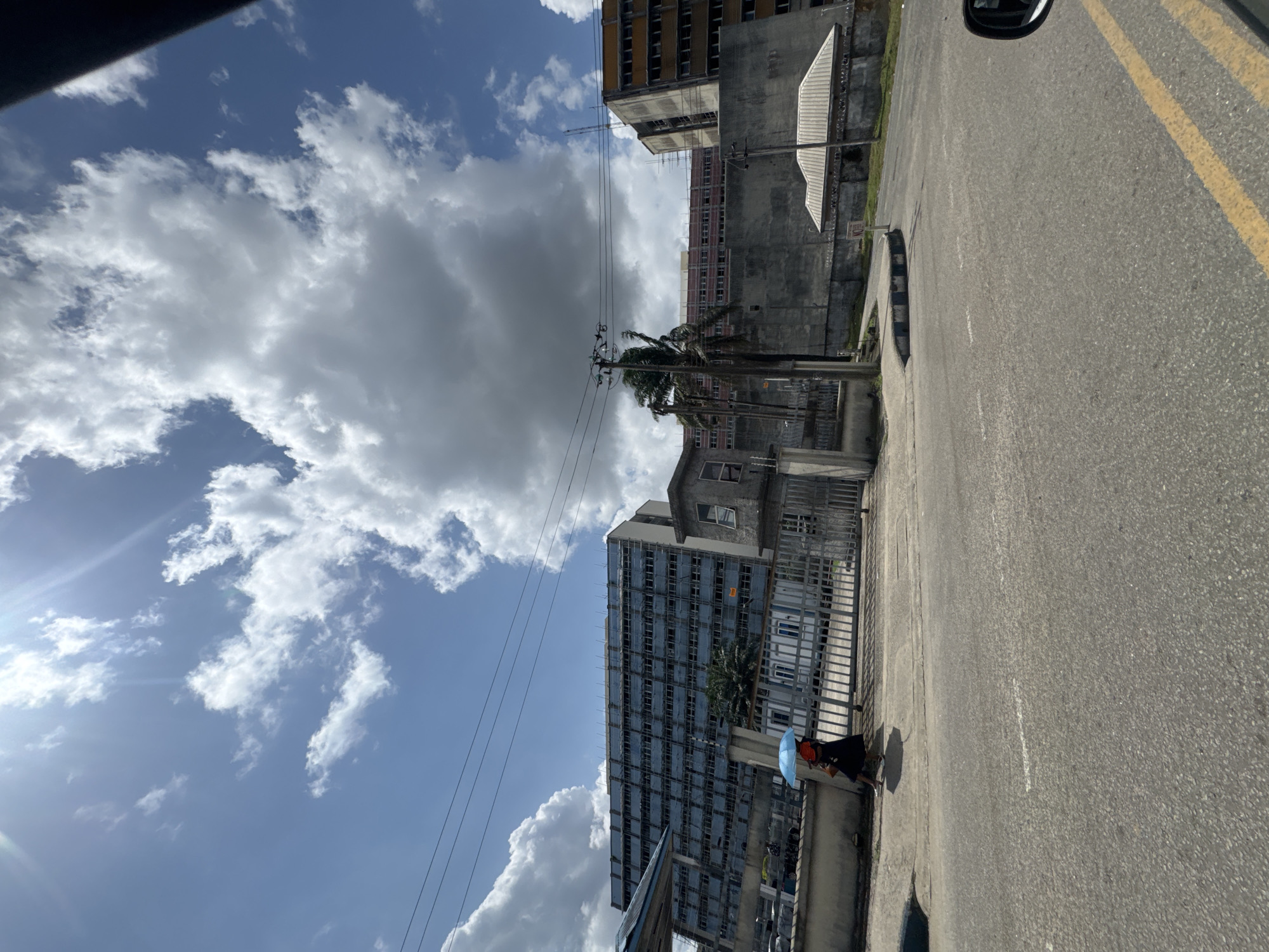 The image shows a street scene in what appears to be Port Harcourt, Nigeria. The angle suggests the photo was taken from a moving vehicle. A woman carrying a blue umbrella walks on the sidewalk next to the street. There are several tall buildings, some in a state of disrepair or ongoing construction. Overhead electrical wires are visible. The sky is bright with many clouds. The road surface appears to be asphalt with yellow lane markings.