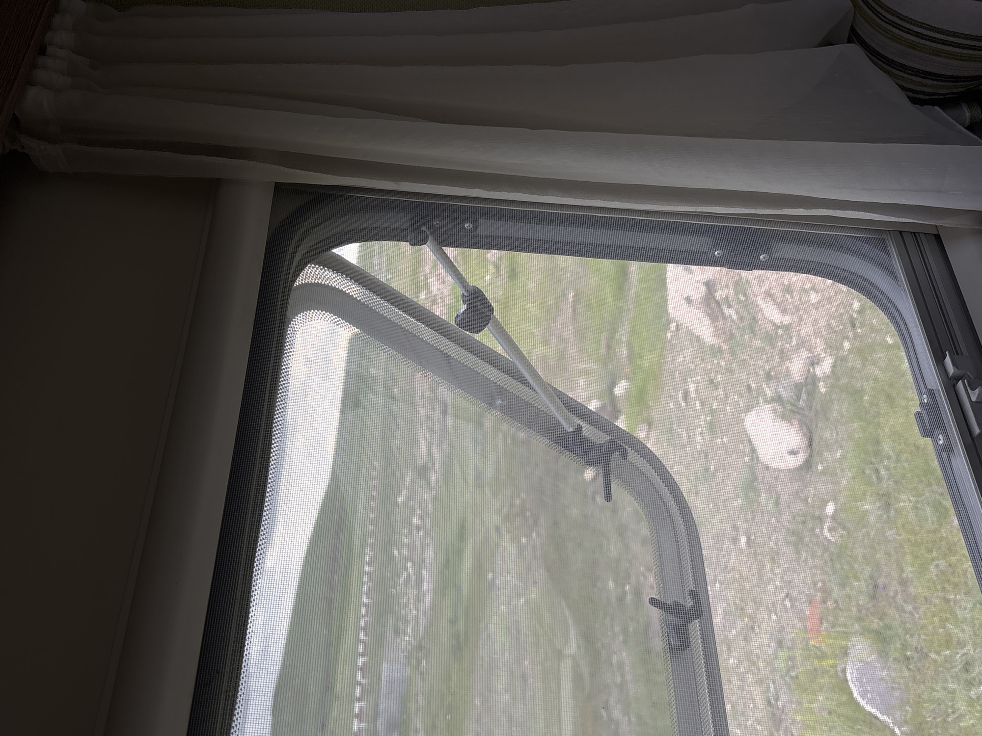 A close-up shot shows a window partially opened with a screen. The view through the screen reveals an outdoor scene in Tlik, Armenia, composed of a grassy field, sparsely dotted with rocks and patches of bare earth. The window is fitted with a mechanism to hold it open at various angles, with a white rod connected to the frame by black fixtures. Curtains are visible at the top of the frame, indicating the window is located inside a structure. The focus is on the window and its immediate surroundings, with the outdoor landscape providing context.