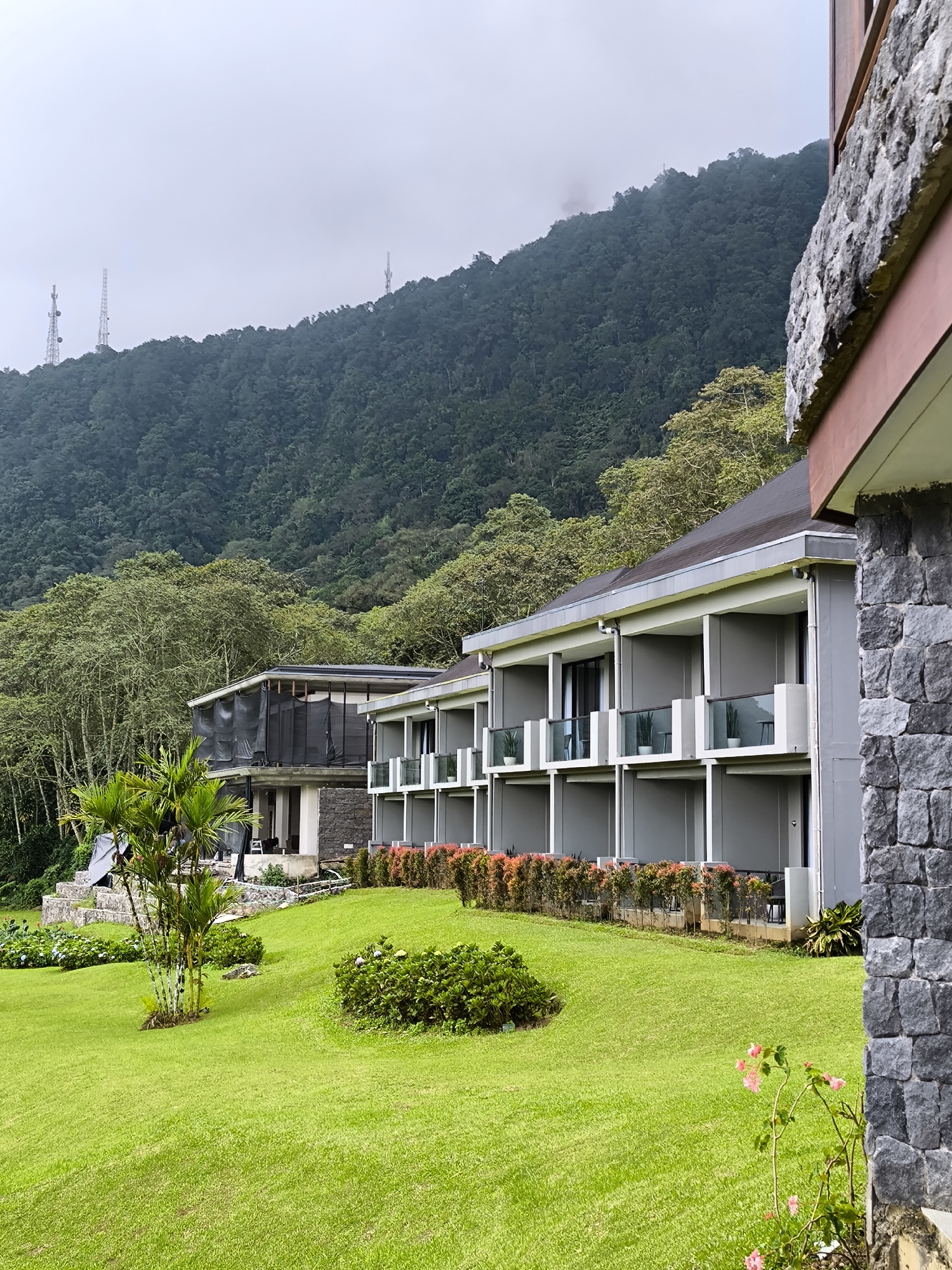 The image depicts a multi-story building complex situated on a manicured lawn in Wanagiri, Indonesia. The gray buildings feature balconies with glass railings and potted plants. A building under construction is visible to the left. Orange and green foliage lines the base of the buildings. A lush green lawn stretches towards a forested mountain in the background. Telecommunication towers are visible on the mountain ridge. The sky is overcast. A stone-covered section of the building is partially visible in the foreground on the right. A small cluster of pink flowers grows in the lower right corner.