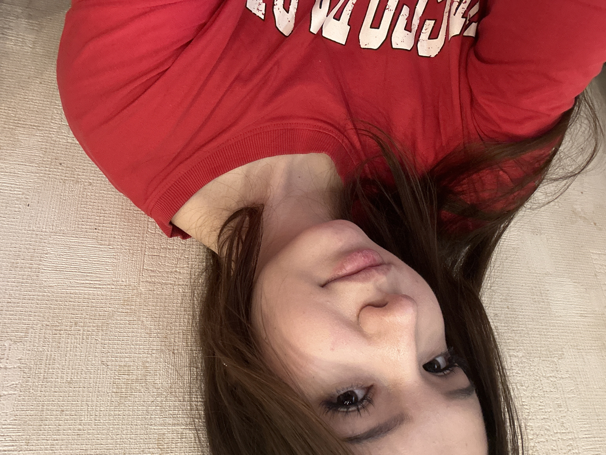 A young adult female is depicted in a close-up photograph, appearing to be a selfie, taken indoors in Naberezhnye Chelny, Russia. The subject has long, dark brown hair, parted on the left side, falling over her shoulders. Her facial features include dark eyes with prominent eyelashes, a small nose, and full lips, with the lower lip exhibiting minor chapping. A subtle, gentle smile is present on her face. She is wearing a red, long-sleeved crew-neck shirt with partially visible white, distressed lettering on the front, with the letters "CROC" discernible. The background consists of an interior wall with a textured, light beige or off-white surface, exhibiting a subtle horizontal pattern, indicative of wallpaper or a similar finish. The lighting is soft and even.