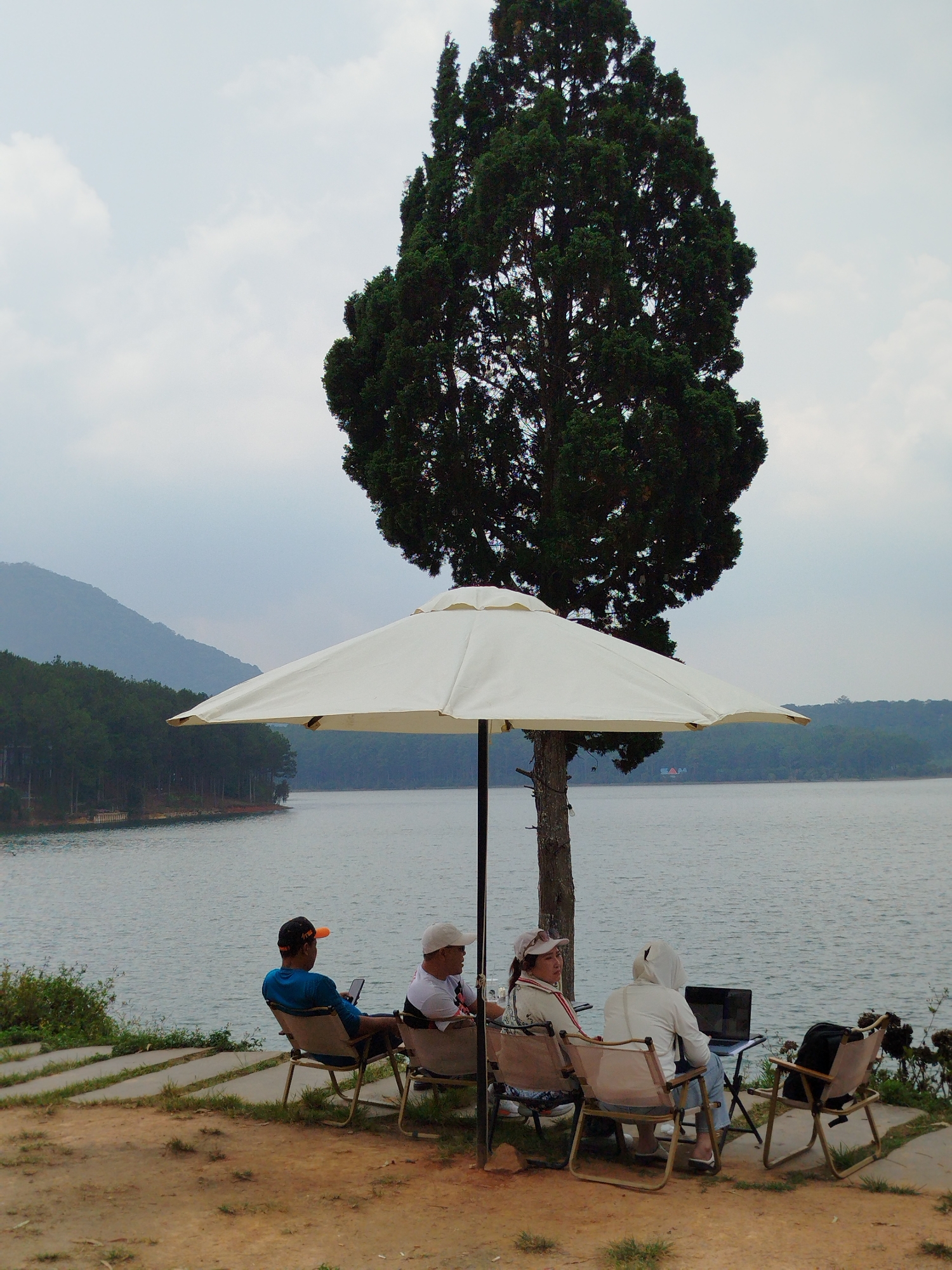 Four people are sitting in chairs under a large white umbrella next to a lake in Da Lat, Vietnam. A tall, dark green tree stands behind the umbrella. The people are positioned facing the lake. The person on the left is wearing a blue shirt and an orange baseball cap. The second person is wearing a white baseball cap and a white shirt. The third person is wearing a hat and has a patterned shirt. The fourth person has a white hooded jacket, blue jeans and slippers. A black laptop is on a small table in front of the fourth person. A black backpack is on the chair to the right. The lake extends to the horizon and is surrounded by green trees and hills. The ground is made of dirt and some grass. The sky is overcast and grey.