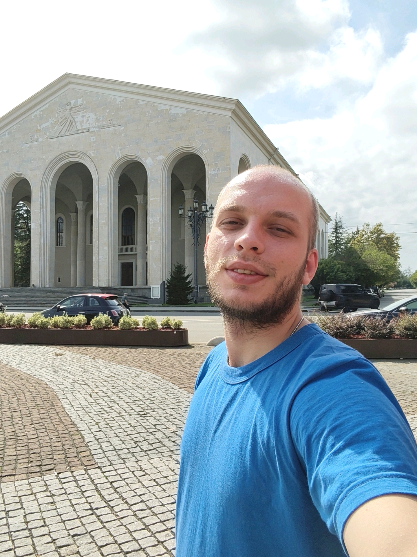 In Ozurgeti, Georgia, a selfie is taken in front of the Ozurgeti Drama Theater. The theater is a large, neoclassical building with a facade featuring multiple arched entrances and columns. A man with a shaved head and beard, wearing a blue t-shirt, is in the foreground, smiling at the camera. Behind him, a black car with a red roof is parked, and a lamppost stands near the entrance of the theater. The ground in front of the theater is paved with cobblestones in a patterned design. The sky is partly cloudy, with visible blue sky and white clouds.