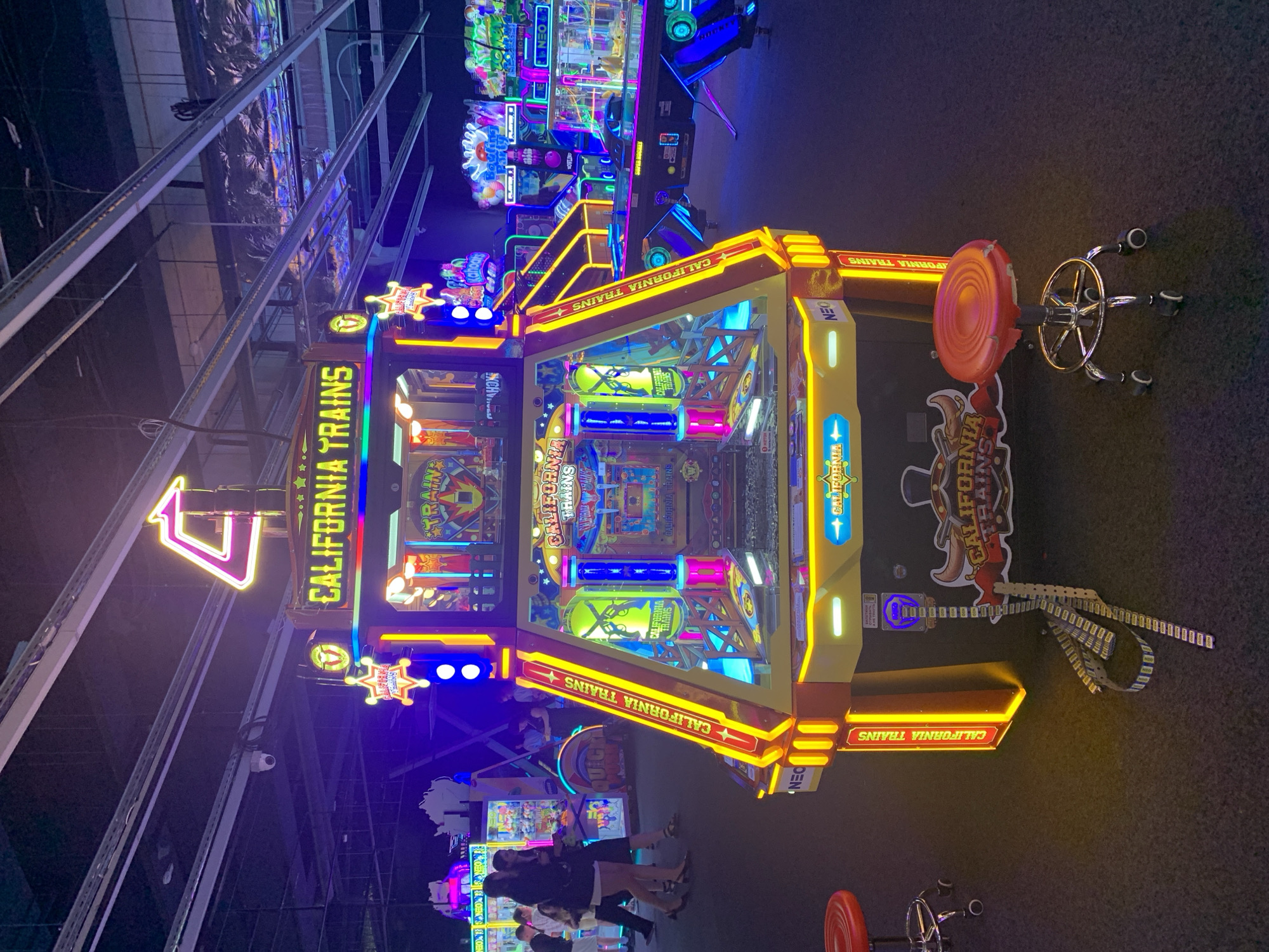 In the dimly lit interior of an arcade, a brightly lit "California Trains" arcade game stands prominently in the foreground. The game features a yellow and red color scheme with neon blue and pink lighting accents. Several people are visible in the background, walking amongst other arcade machines, some of which also have vibrant lighting. The floor is dark carpet, and the ceiling has exposed industrial elements with fluorescent lighting and hanging wires. The overall scene suggests a typical arcade environment with various games and patrons. There is no information in the image to indicate a specific location in the city of Hrodna, Belarus.
