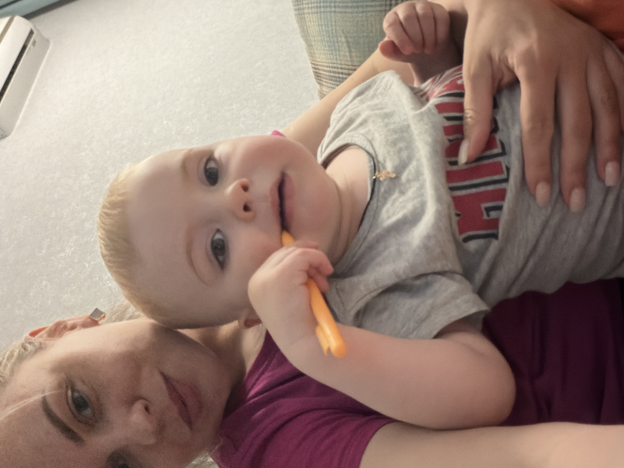 An intimate, high-angle selfie captured indoors in Votkinsk, Russia, features a woman and a young child. The fair-haired child, appearing to be a toddler or baby, is central in the frame, looking directly towards the camera with a curious expression. The child wears a light gray t-shirt with partially visible red and white lettering that seems to spell "HILL" or similar. An orange, cylindrical object, possibly a teether or baby toothbrush, is held in the child's right hand and is near or in their mouth. A small golden pendant hangs from the child's neck.
The woman, with blonde hair and light makeup, is visible on the left side of the frame, positioned below the child. She is wearing a vibrant purple top. Her hand is gently resting on the child's chest, near the t-shirt lettering, displaying light-colored manicured nails.
The background shows a light-colored, textured wall. An air conditioning unit is mounted on the wall in the upper left corner. A plaid or checkered pillow with green and blue tones is visible behind the child's head and upper body, suggesting a comfortable, domestic setting. The overall mood of the image is relaxed and affectionate, capturing a tender moment between the two subjects.