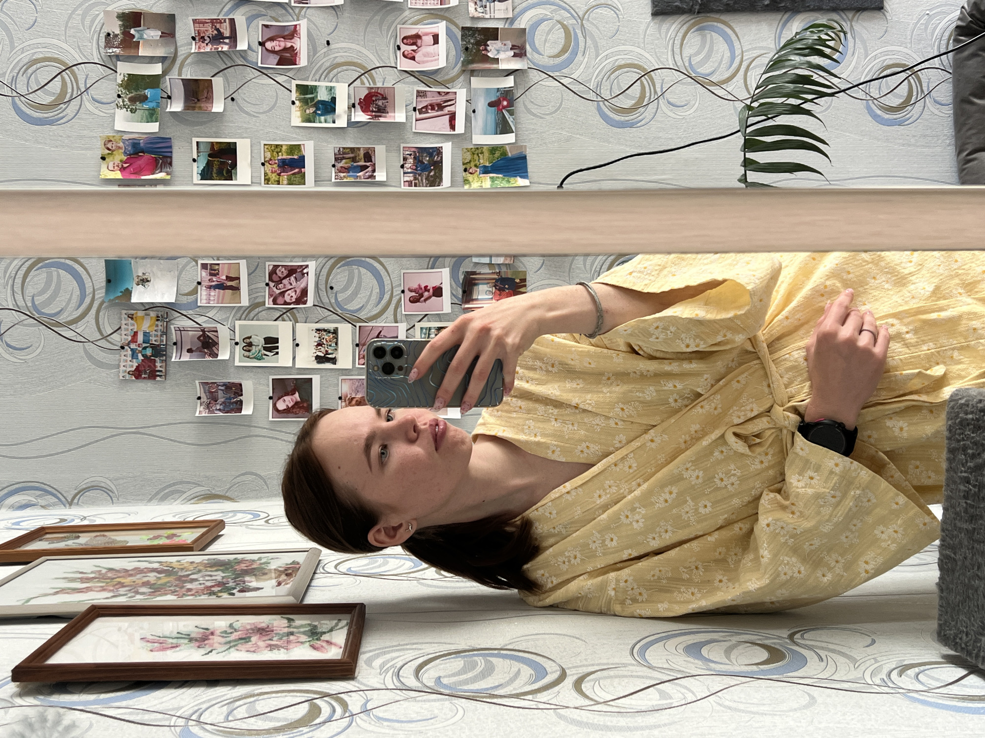 A young woman lies on a patterned surface taking a selfie with a grey smartphone. She is wearing a yellow floral dress and a smartwatch on her left wrist. Behind her, three framed floral embroideries lie on the same patterned surface. In the background, a wall is decorated with a patterned wallpaper and a collection of small photographs attached with black wires. A shelf runs horizontally across the wall, with a green plant and a grey cushion visible above it. The location is likely indoors, in an apartment or house, but the city of Chaikovsky, Russia, cannot be confirmed from the image alone.