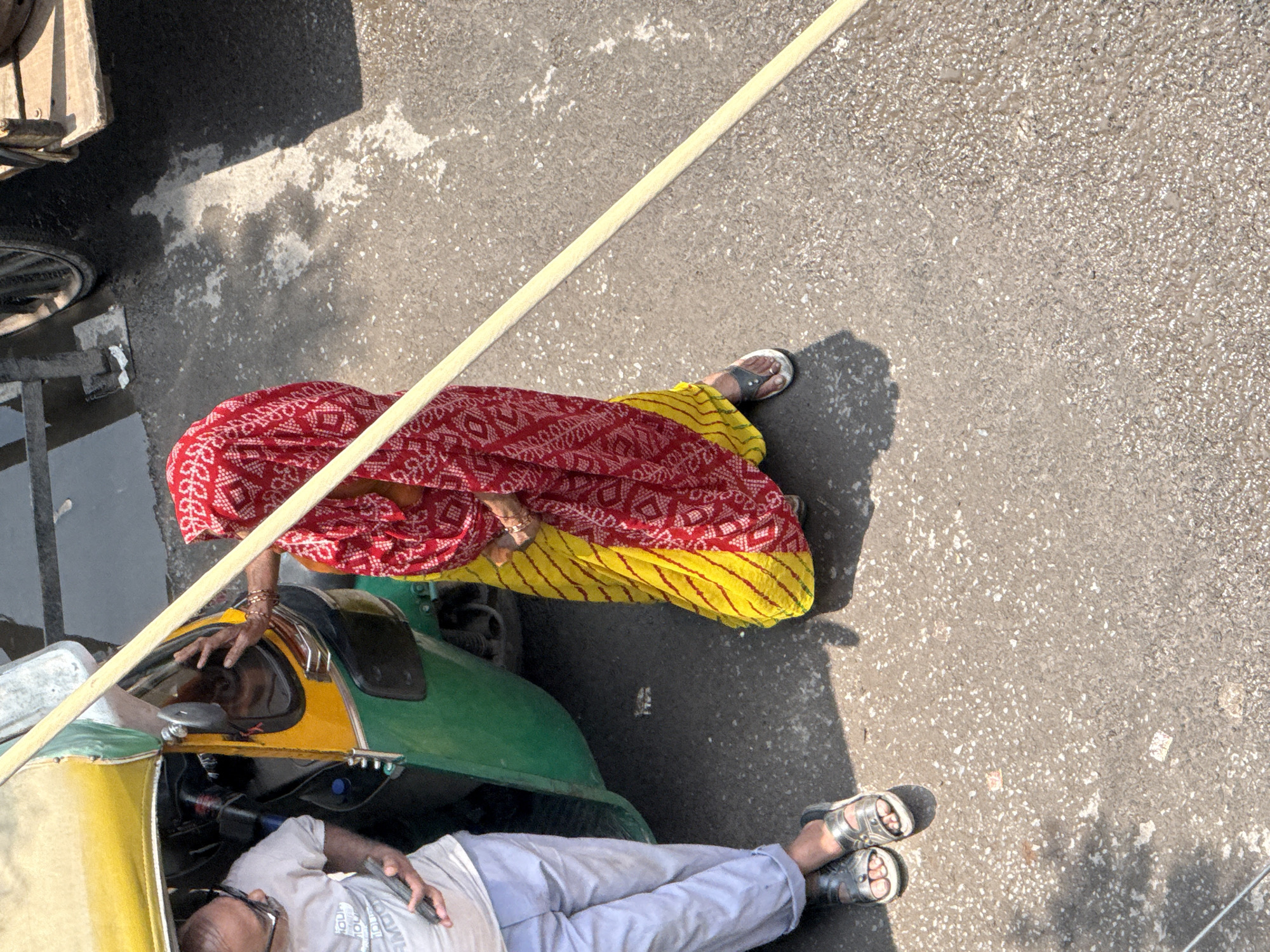 The image shows a street scene in Delhi, India, with an auto-rickshaw as the central element. A man wearing a white t-shirt and light-colored trousers stands next to the open door of the auto-rickshaw, holding a cell phone. He is wearing silver sandals. To his right, a woman is dressed in a red and white patterned scarf and a yellow sari. She is partially obscured by the auto-rickshaw and is holding a long, thin wooden pole that extends diagonally across the frame. Her left hand is raised, appearing to be in contact with the auto-rickshaw. The ground is paved with asphalt and shows patches of dampness and scattered debris. In the background, parts of a cart with a large wheel are visible. The lighting suggests it is daytime, with strong sunlight casting shadows.