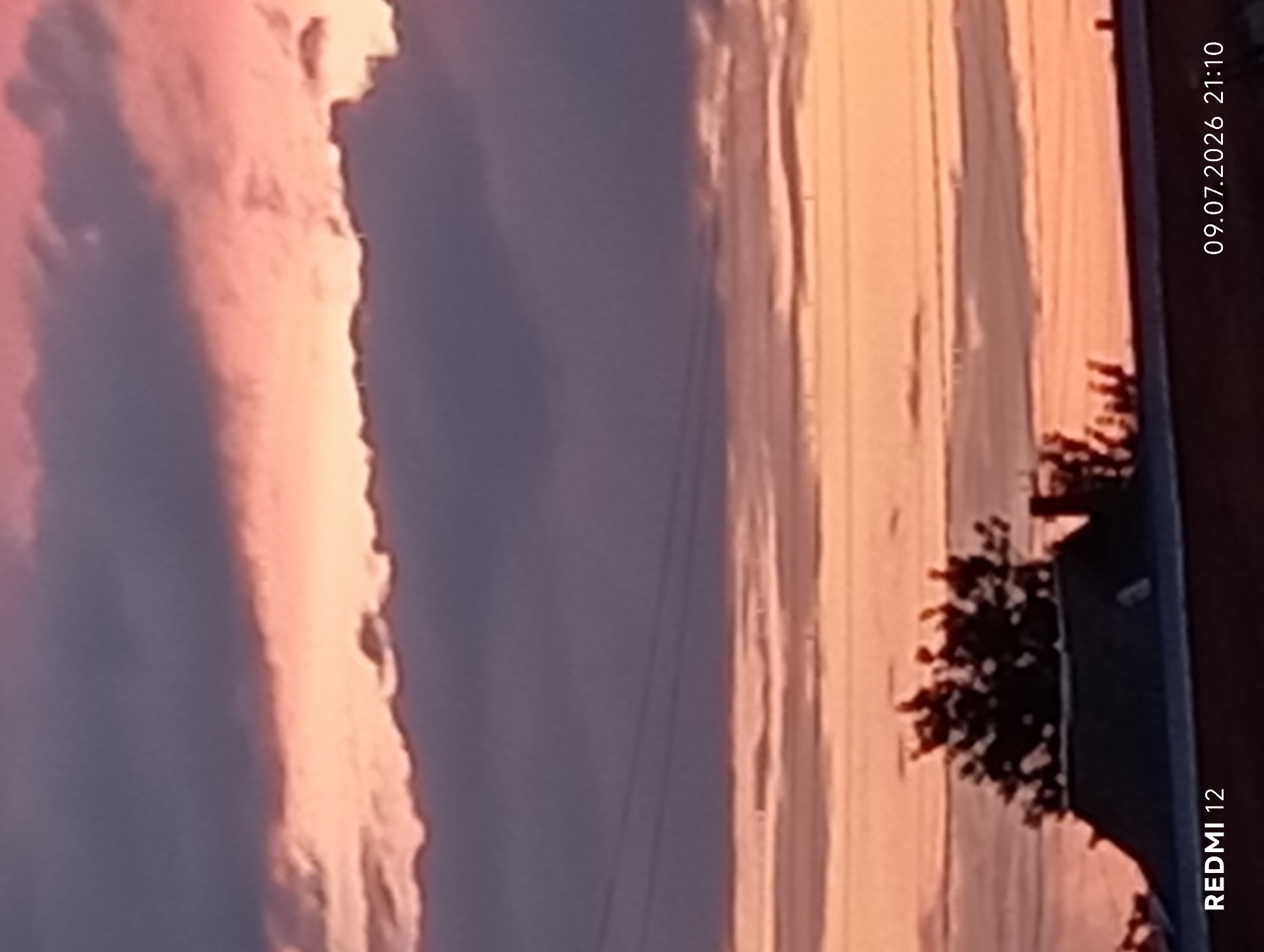 The image depicts a sunset over the city of Smila, Ukraine. The sky is filled with dramatic clouds, tinged with shades of pink, orange, and purple. Power lines are visible across the sky, indicating an urban or suburban setting. In the foreground, the silhouette of a building with a dark roof is visible, topped by trees and a chimney. The bottom left corner of the image contains the text "REDMI 12", and the bottom right corner displays "09.07.2026 21:10", suggesting this is a timestamp from a Redmi 12 mobile device. The scene is peaceful and atmospheric, capturing the natural beauty of a sunset in a populated area.
